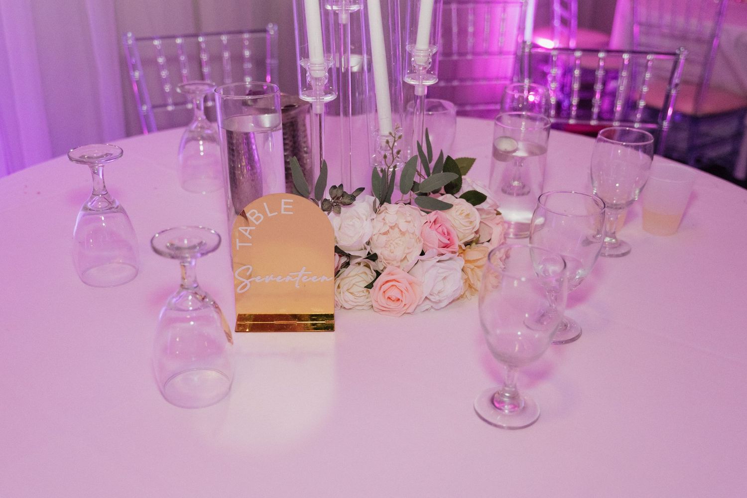 Purple-lit reception tables with elegant place settings and glassware.