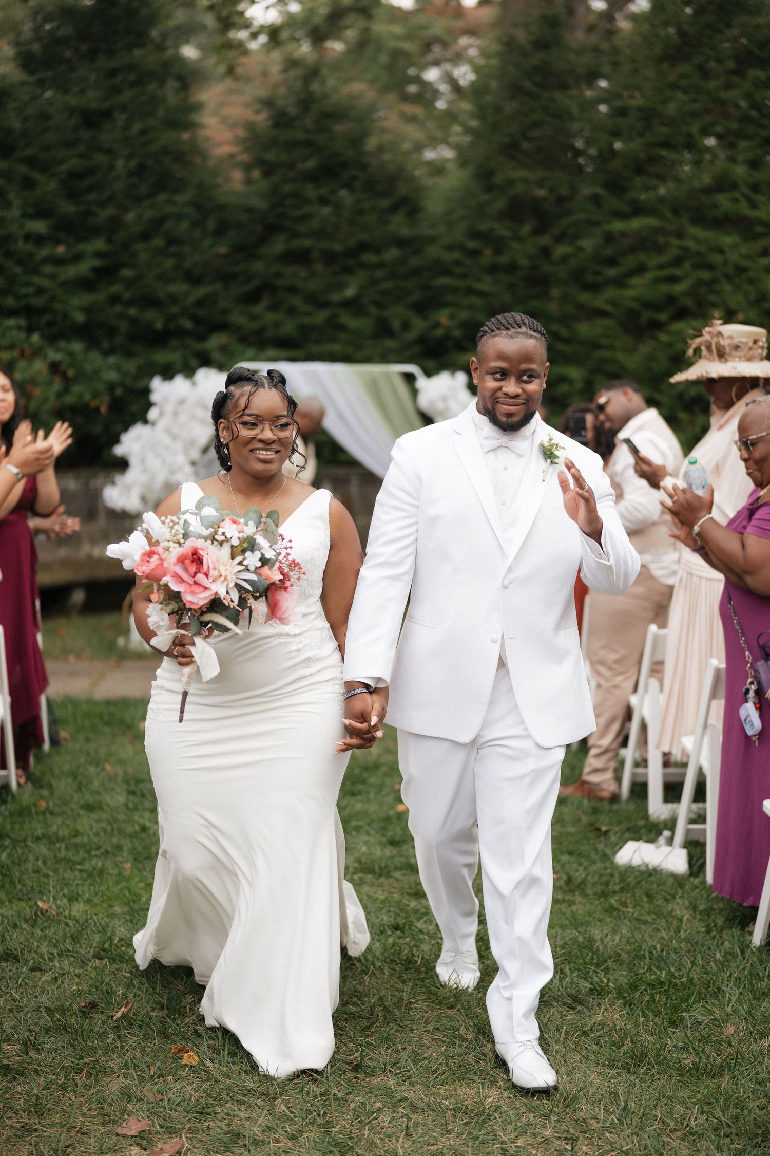 Wedding party celebrates joyfully while walking on grass in white attire and purple bridesmaid dresses.