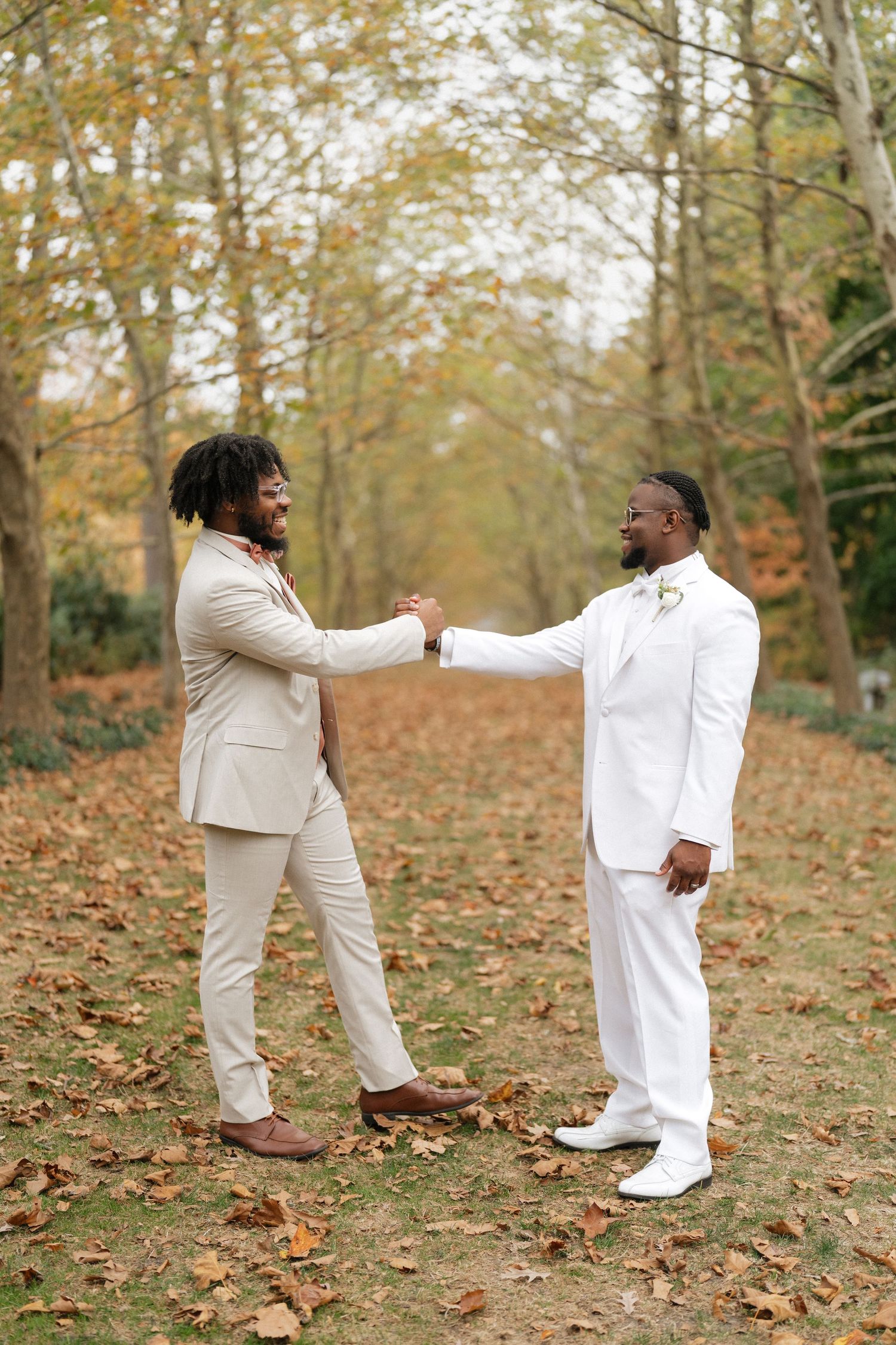 Two people in light colored suits greet each other warmly among autumn trees with fallen leaves.