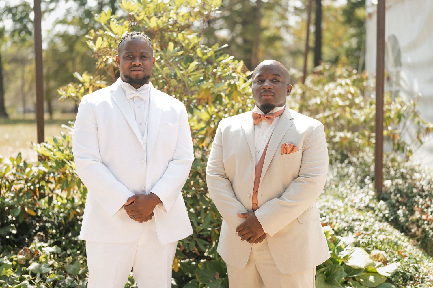 Two people in white formal attire stand together in a garden setting with sunlit foliage in the background.