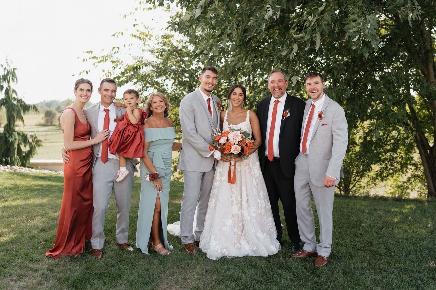 Large wedding party in red and gray attire poses together on grassy hillside.
