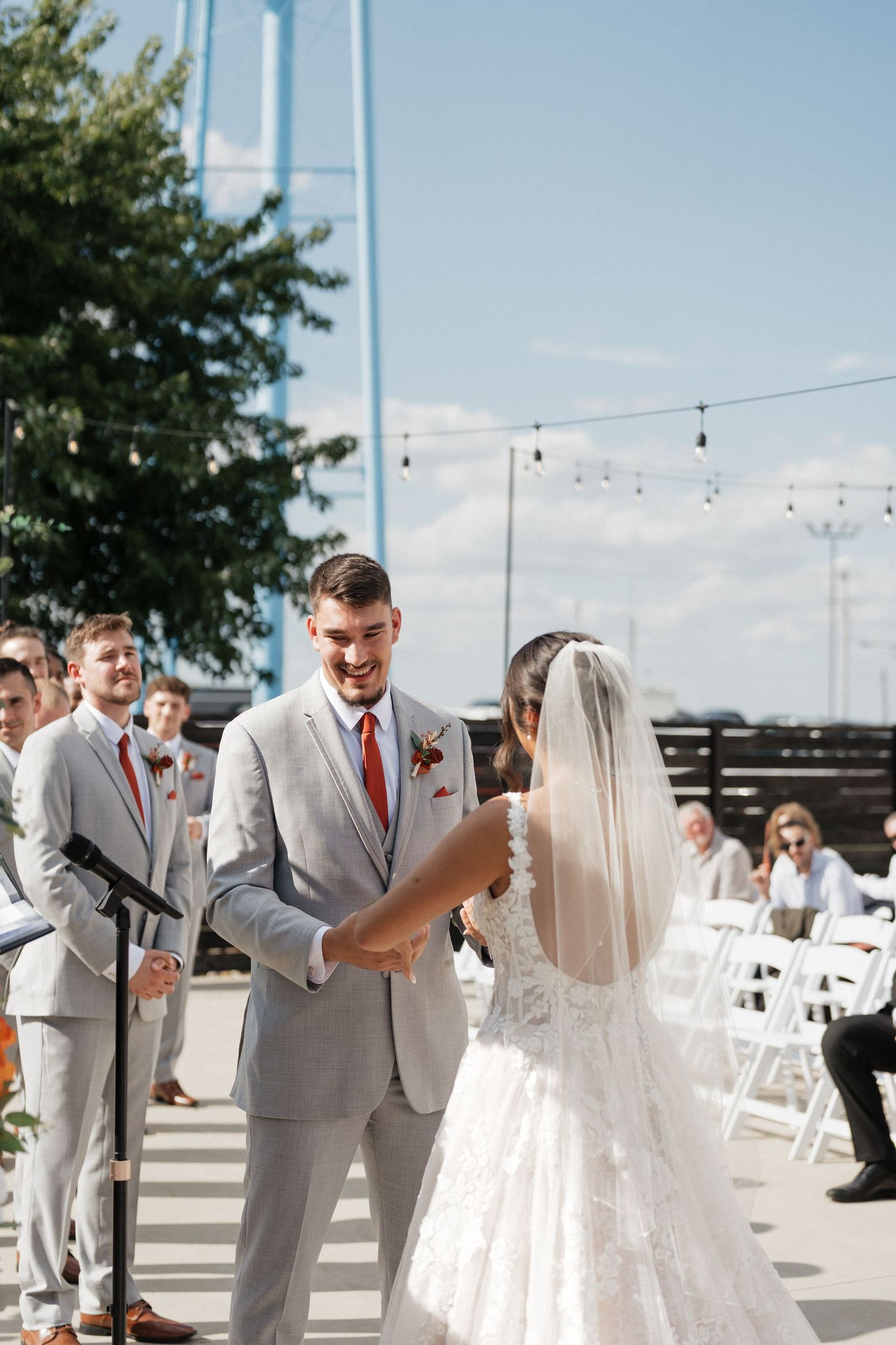 A couple exchanges vows during an outdoor wedding ceremony with string lights and blue sky overhead.