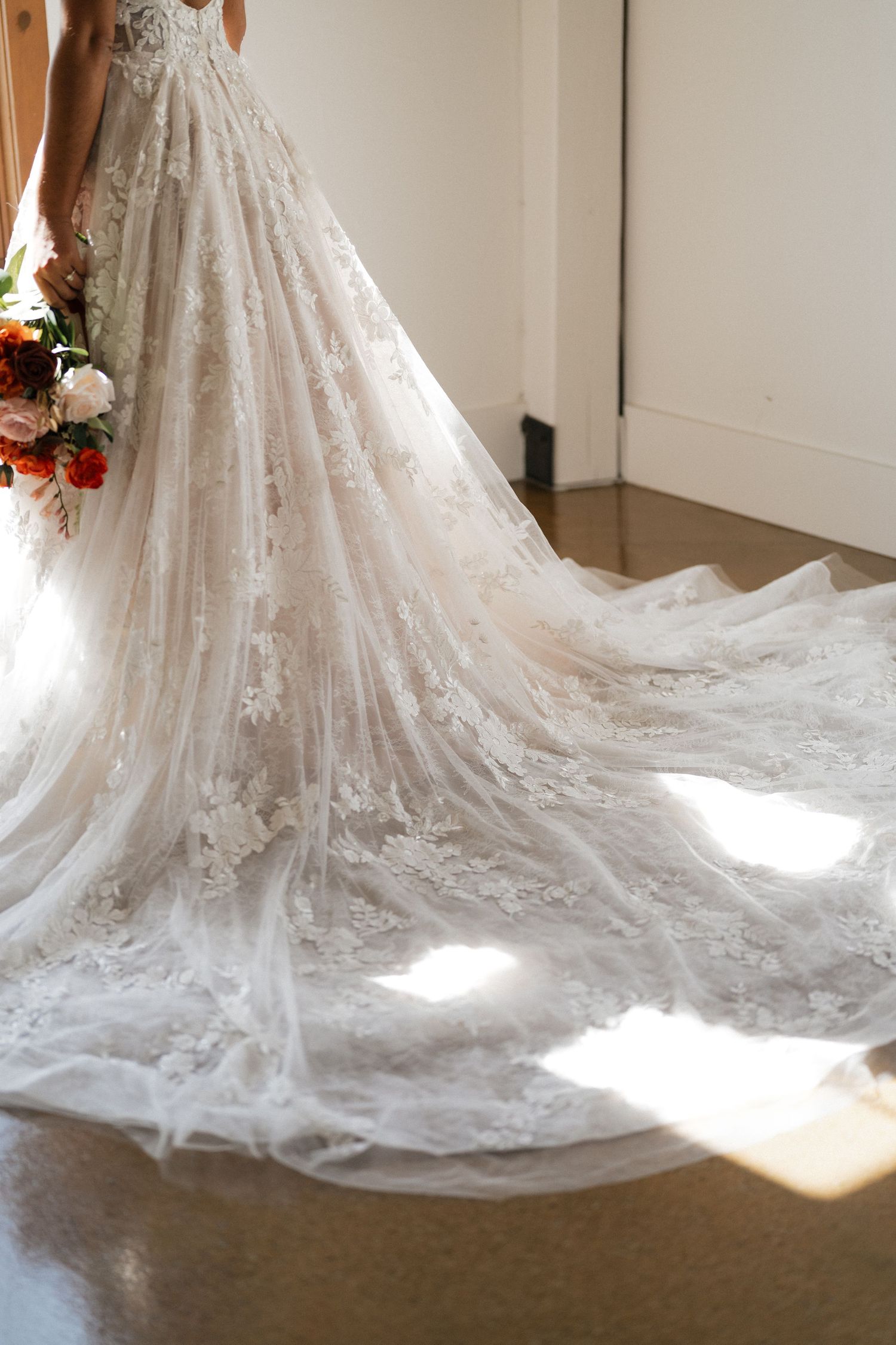 Detailed view of an ornate lace wedding dress train flowing across a wooden floor in natural light.
