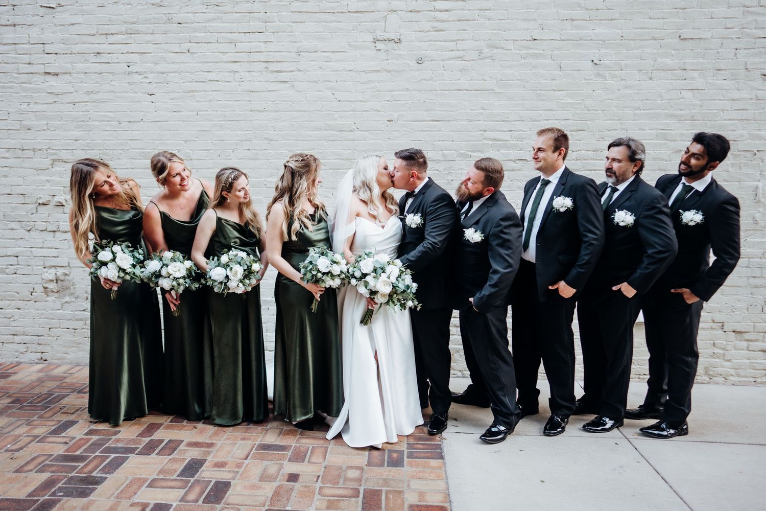 Wedding party poses for group photos against white brick wall in formal attire.