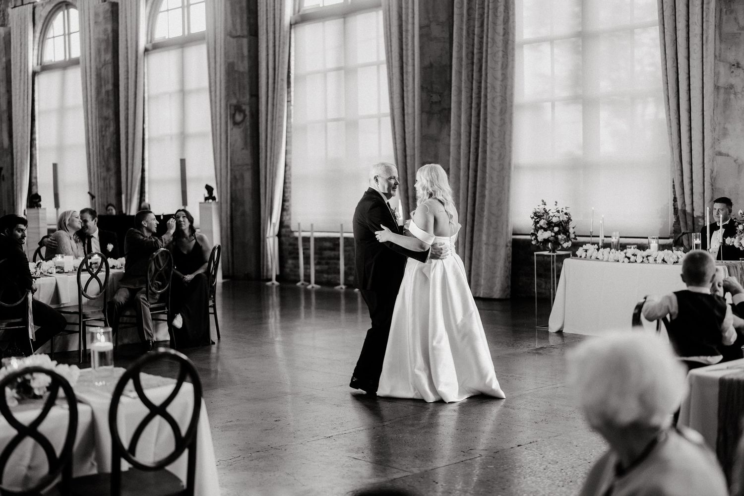 Wedding couple dances in an elegant room with large windows and flowing curtains.