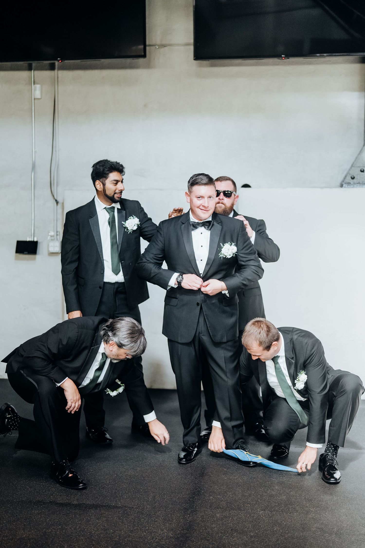 Groomsmen in black tuxedos pose playfully in a modern venue space.