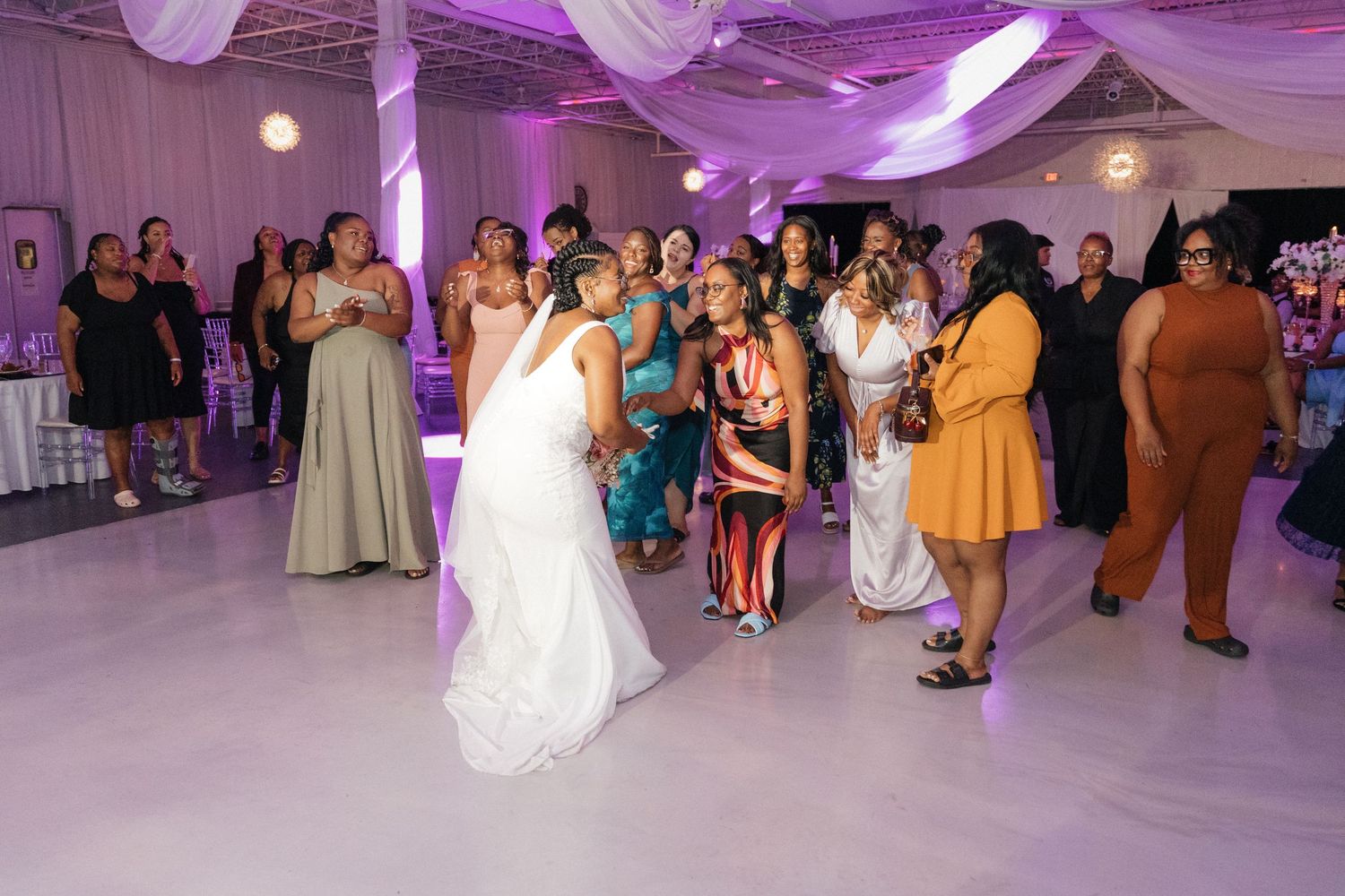 Wedding reception dance floor filled with guests under purple lighting and white draping.