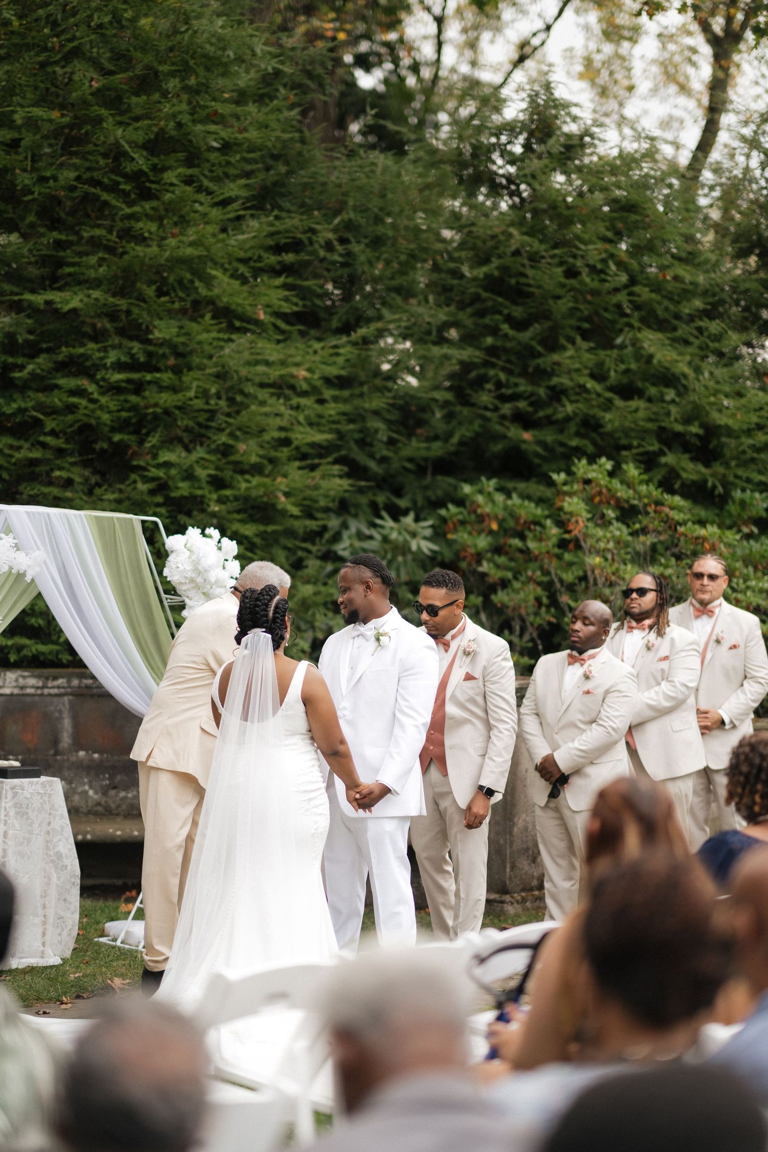 Outdoor wedding ceremony setup with white draping and guests seated beneath tall evergreen trees.