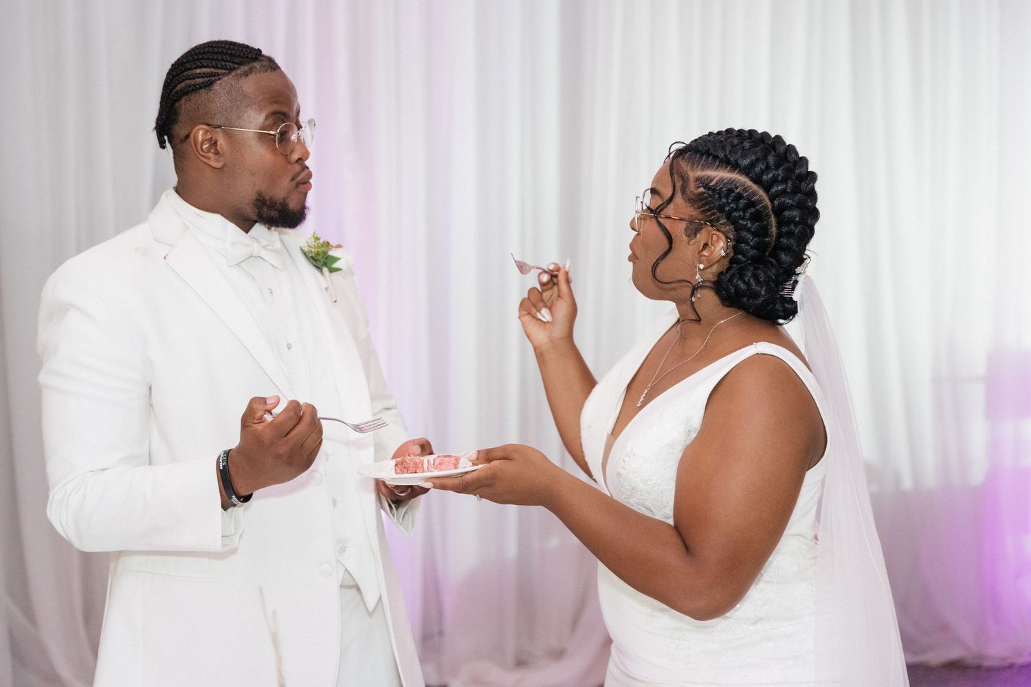 A couple in white wedding attire share cake during their reception celebration.