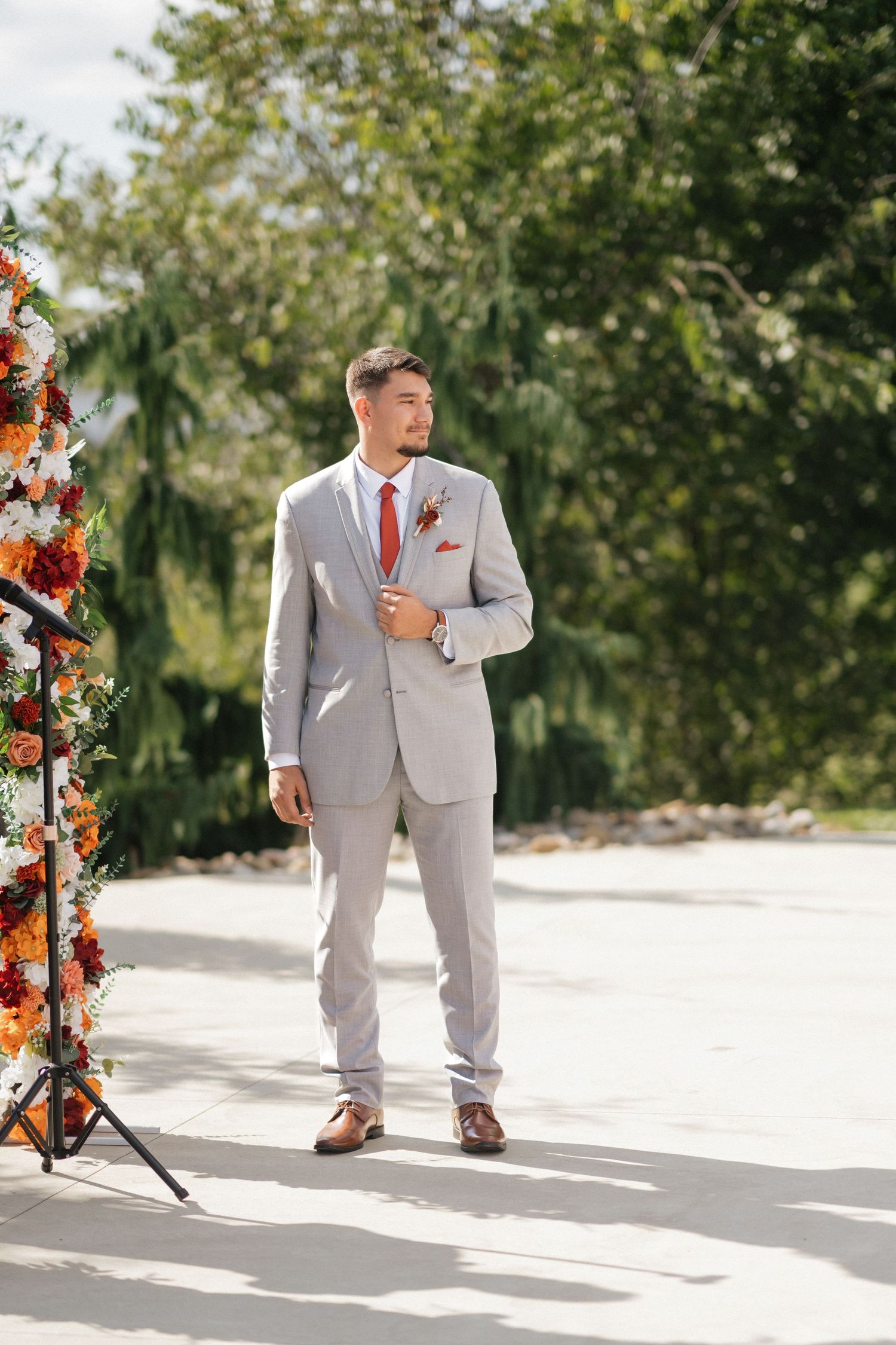 Groom in light grey suit stands in sunlight next to floral wedding arch decorations.