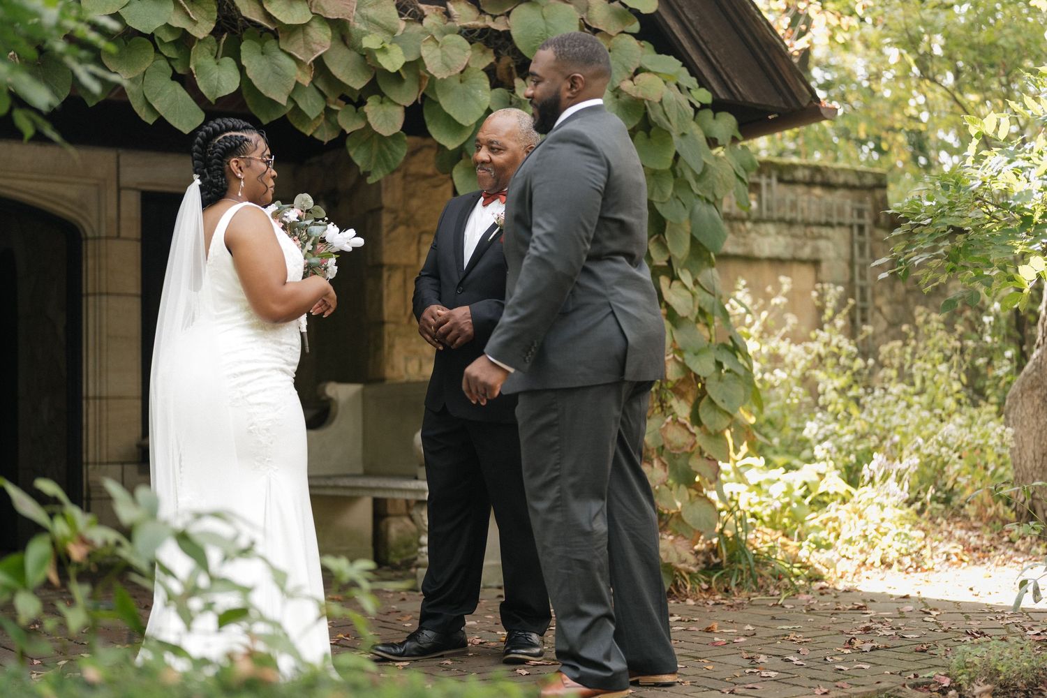 Outdoor wedding ceremony taking place in a lush garden setting with stone architecture.