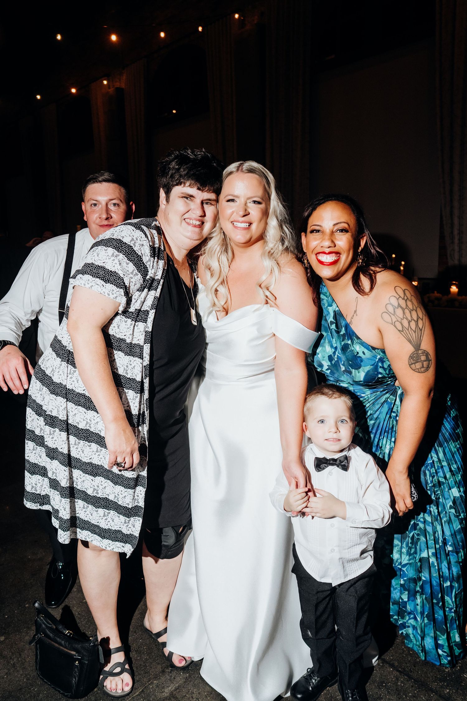 Wedding party poses together for fun group photo at reception venue with string lights in background.