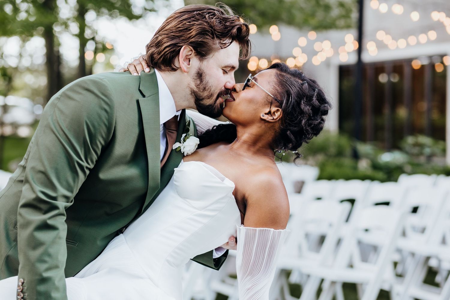 Romantic wedding couple embraces during outdoor ceremony with white chairs and string lights.