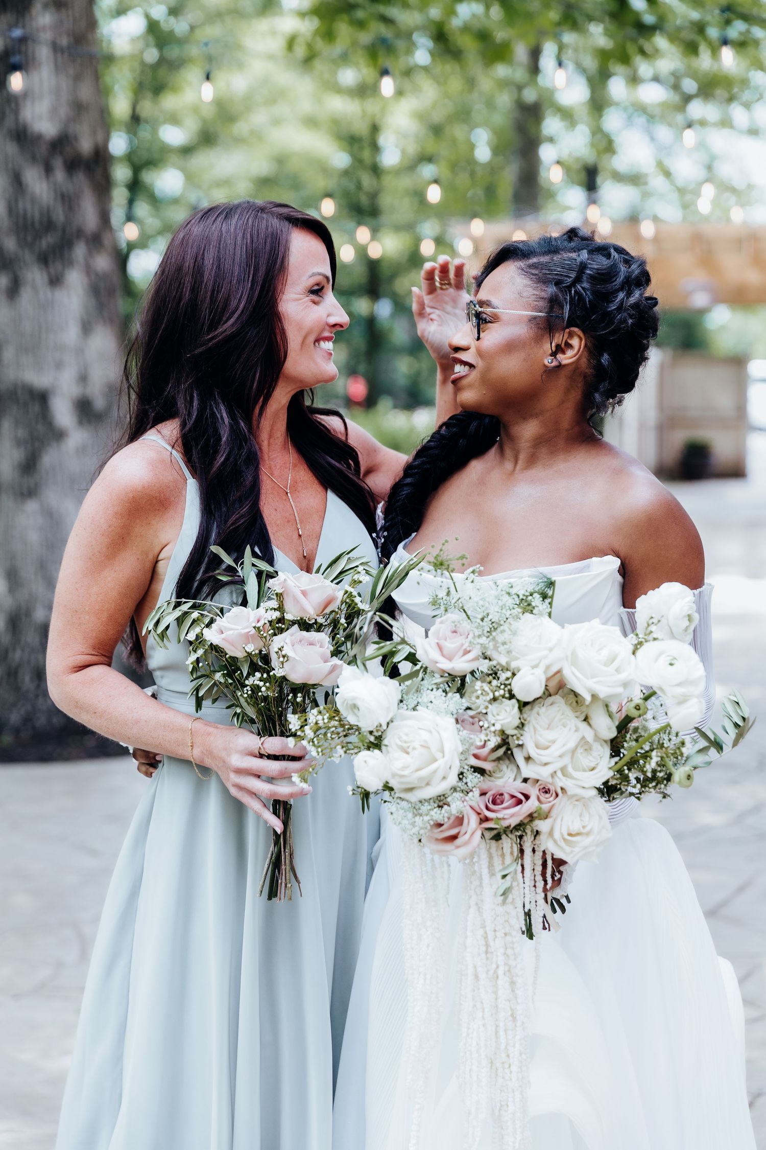 Two people share a joyful moment while holding elegant white rose bouquets at an outdoor wedding.