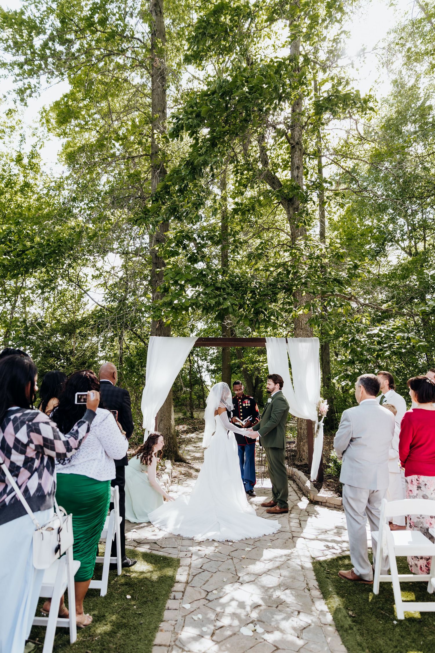 Outdoor wedding ceremony setup with white chairs and fabric drapes among trees.