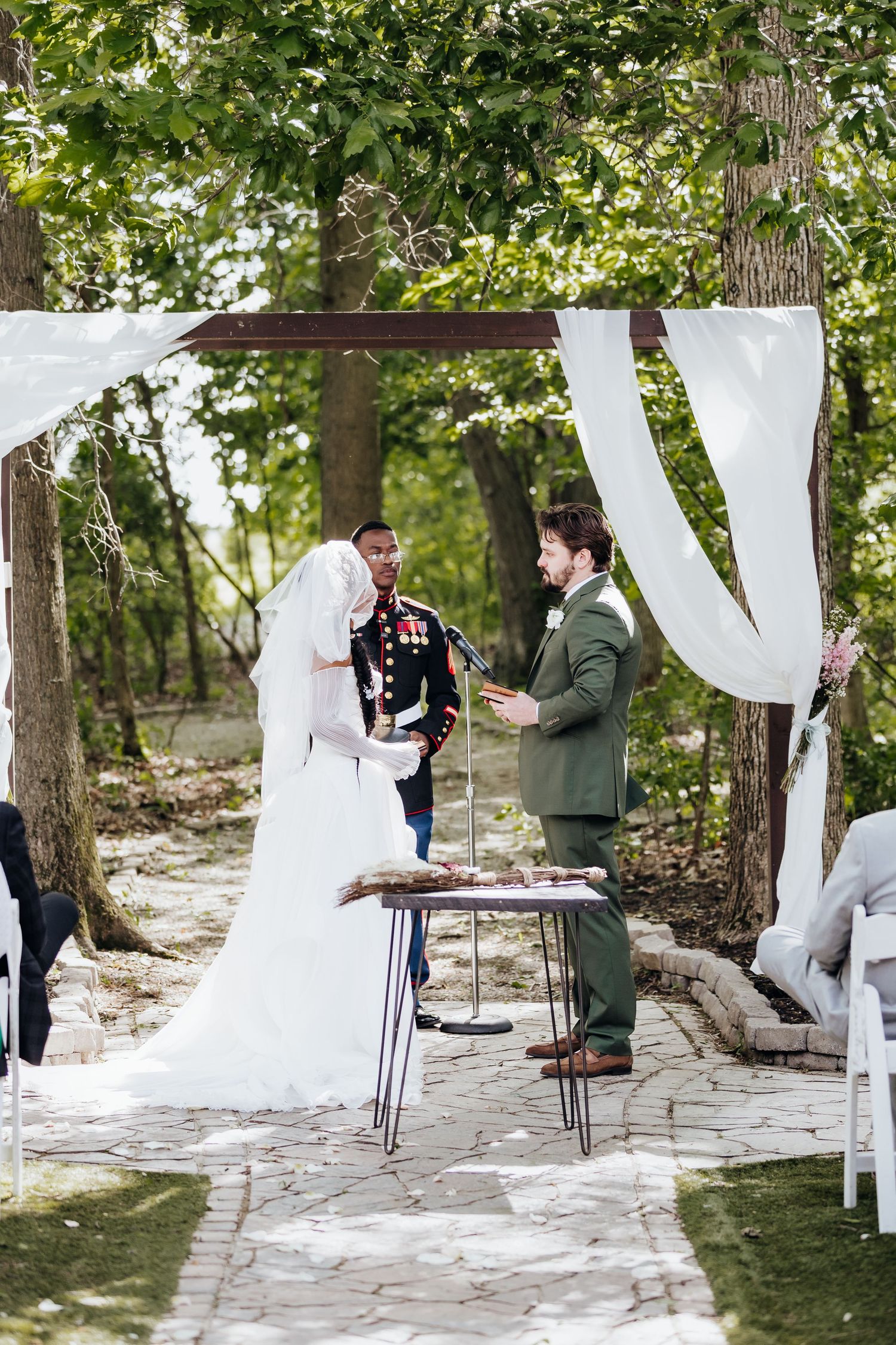 Wedding couple shares a special moment during their first look ceremony on a stone path surrounded by trees and white drapes.