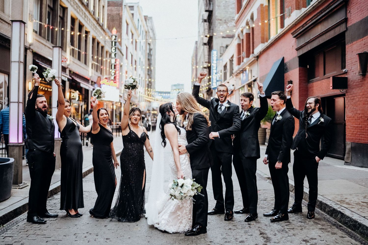 Wedding party in black attire poses on a charming city street with string lights overhead.