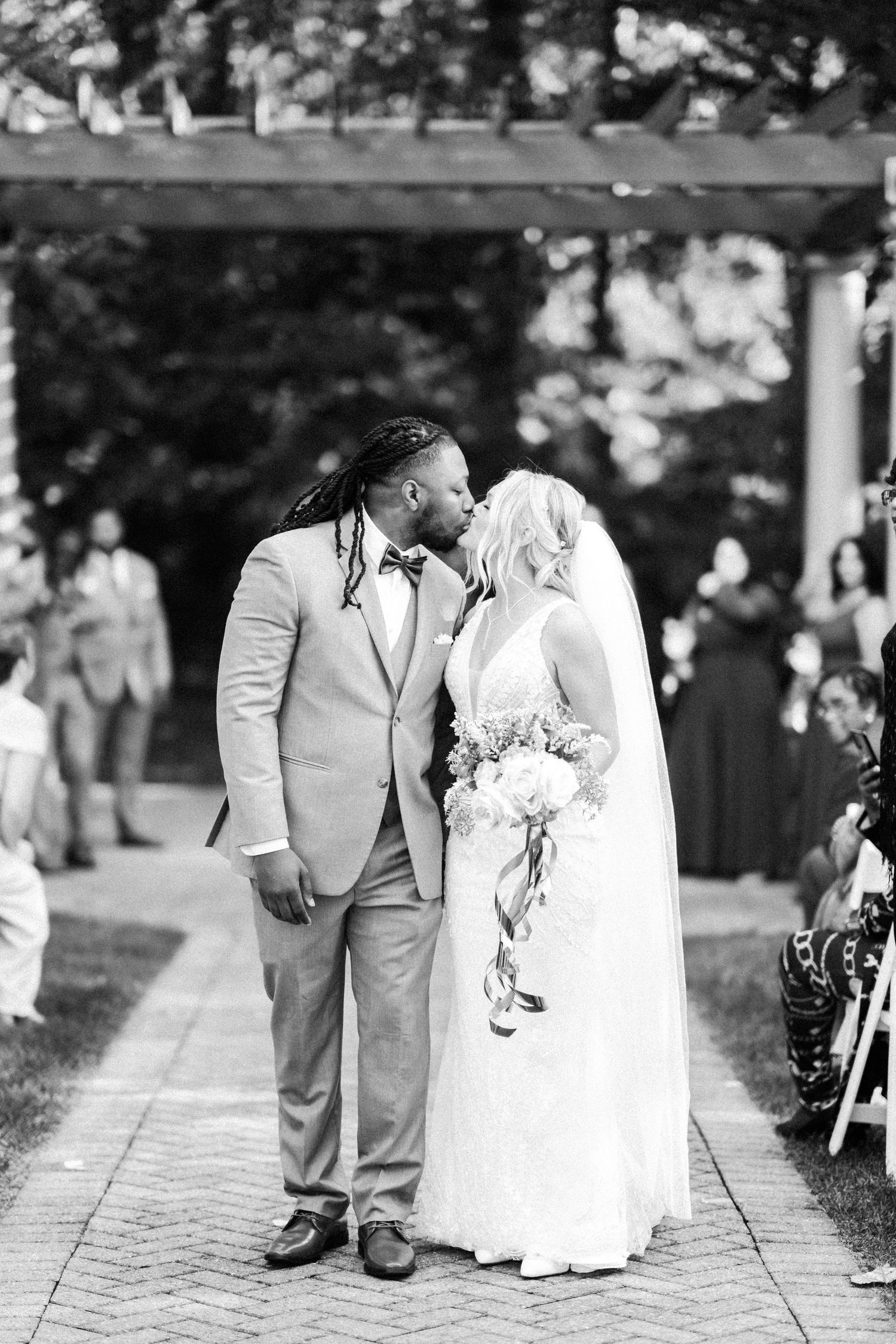 A couple shares their first kiss during an outdoor wedding ceremony under a wooden arch.
