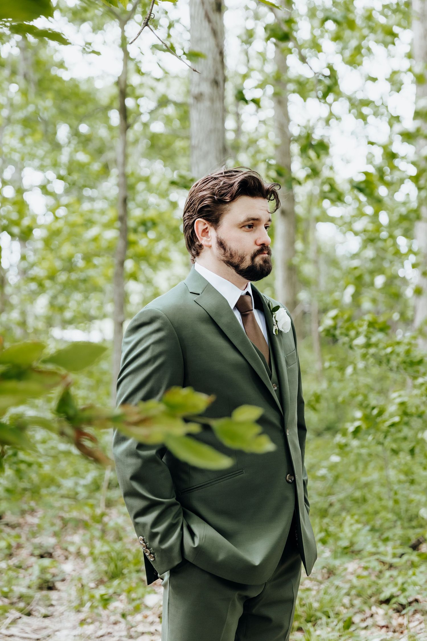 Groom in dark green suit poses thoughtfully in a wooded outdoor setting.