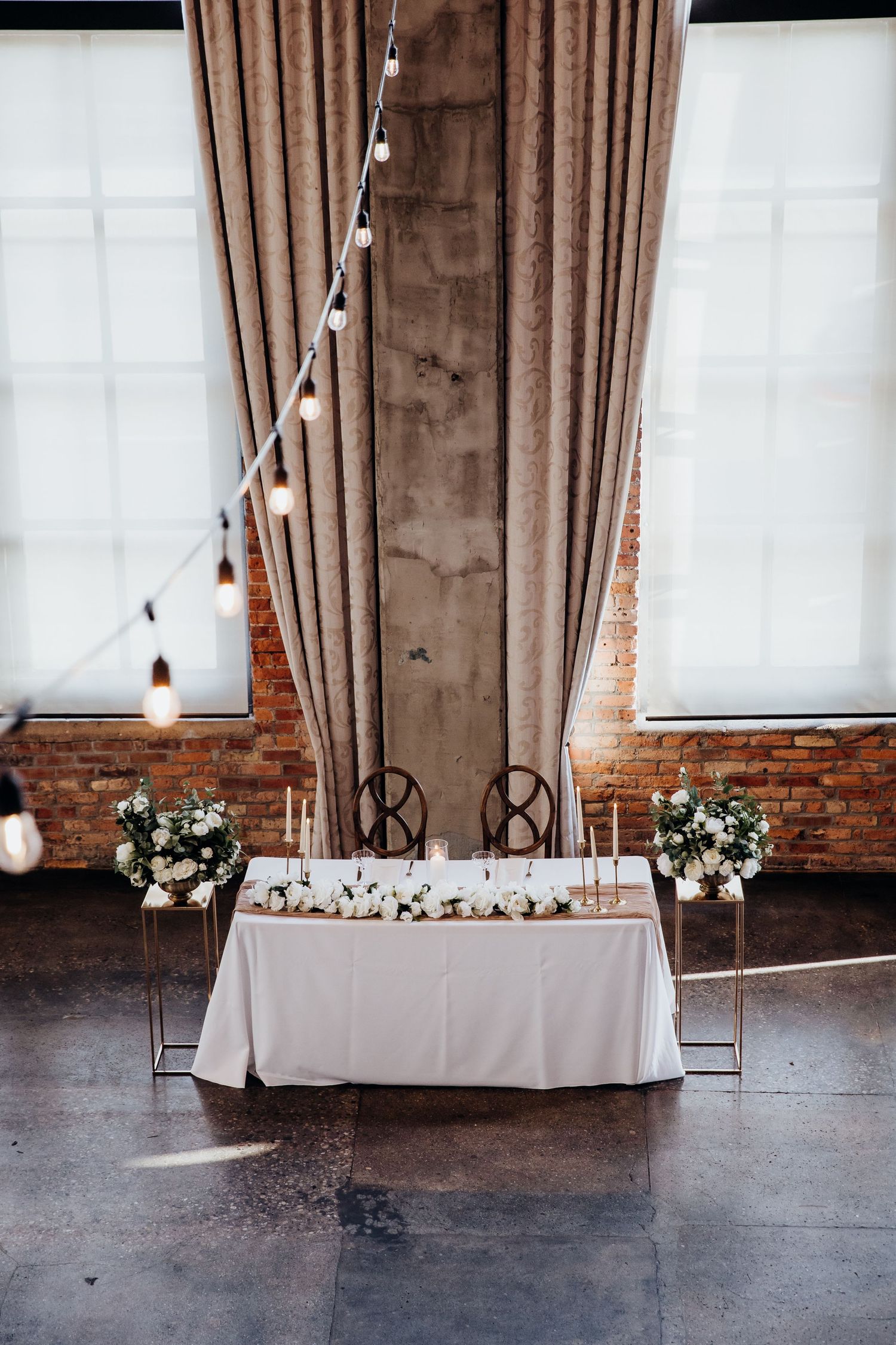 Reception tables with white linens and floral centerpieces set against exposed brick walls and draped windows.