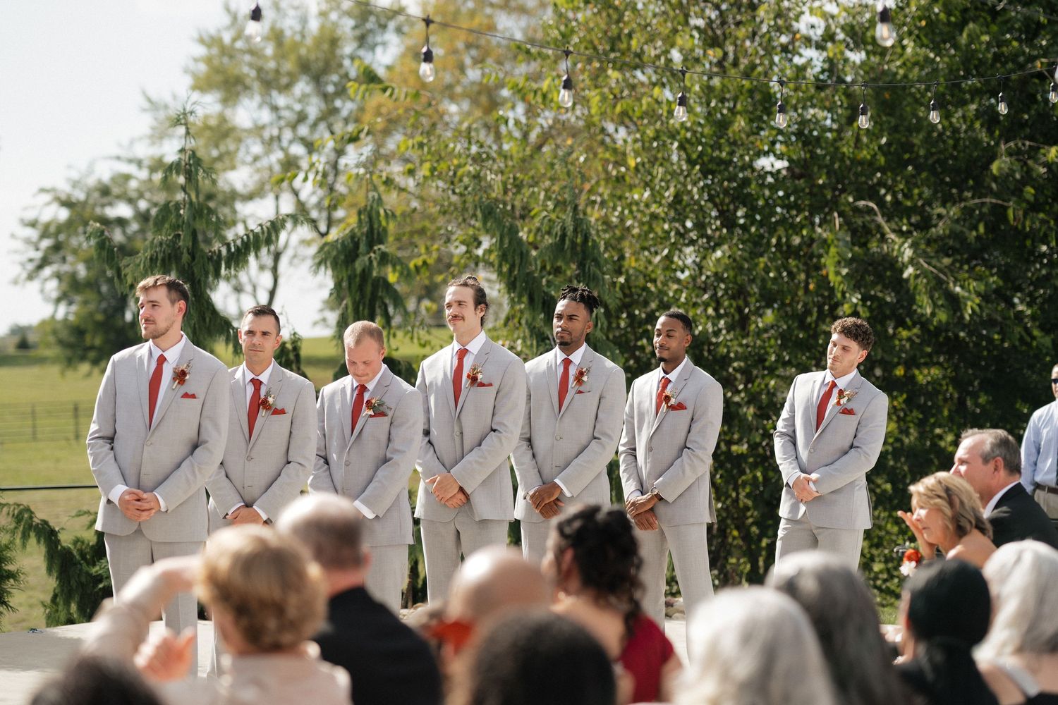 Wedding party in light gray suits and coral accents stands in line during an outdoor ceremony.