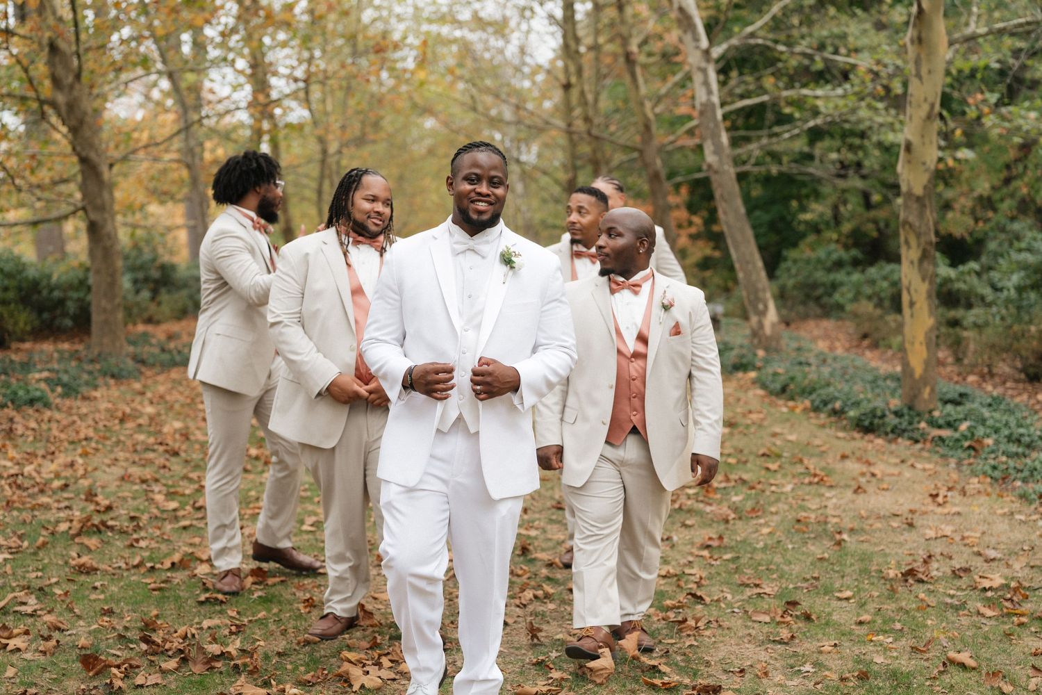 Wedding party in all-white attire walks through autumn garden setting.