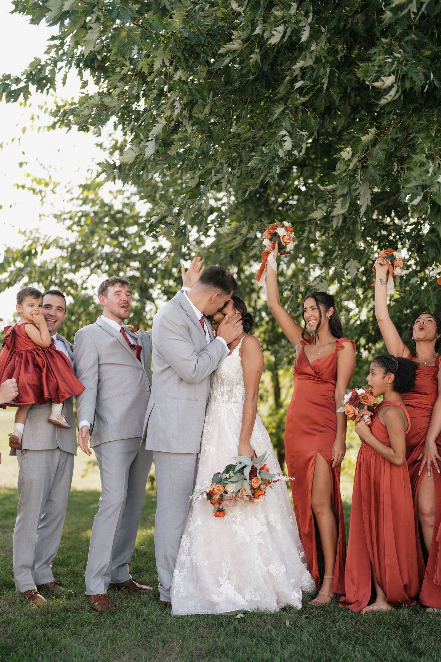 Wedding party poses outdoors wearing gray suits and rust-colored dresses under green trees.
