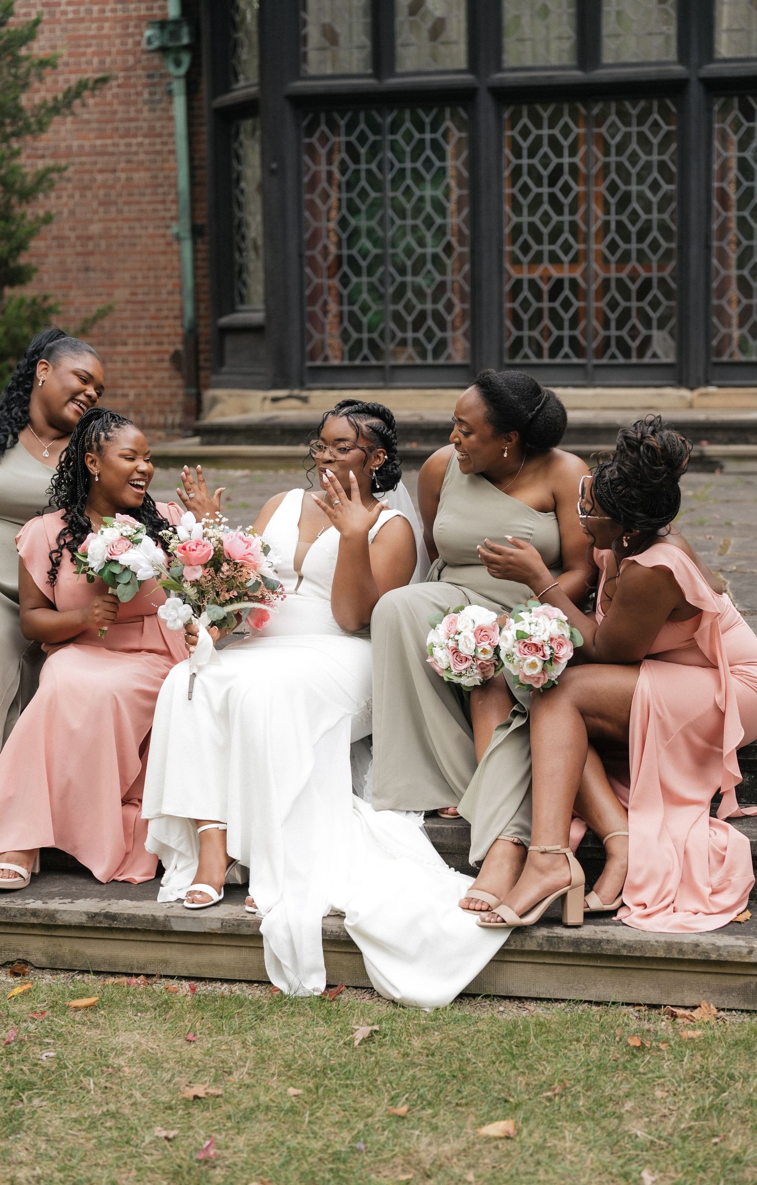 Wedding party in pink dresses relaxing on stone steps of a historic brick building.