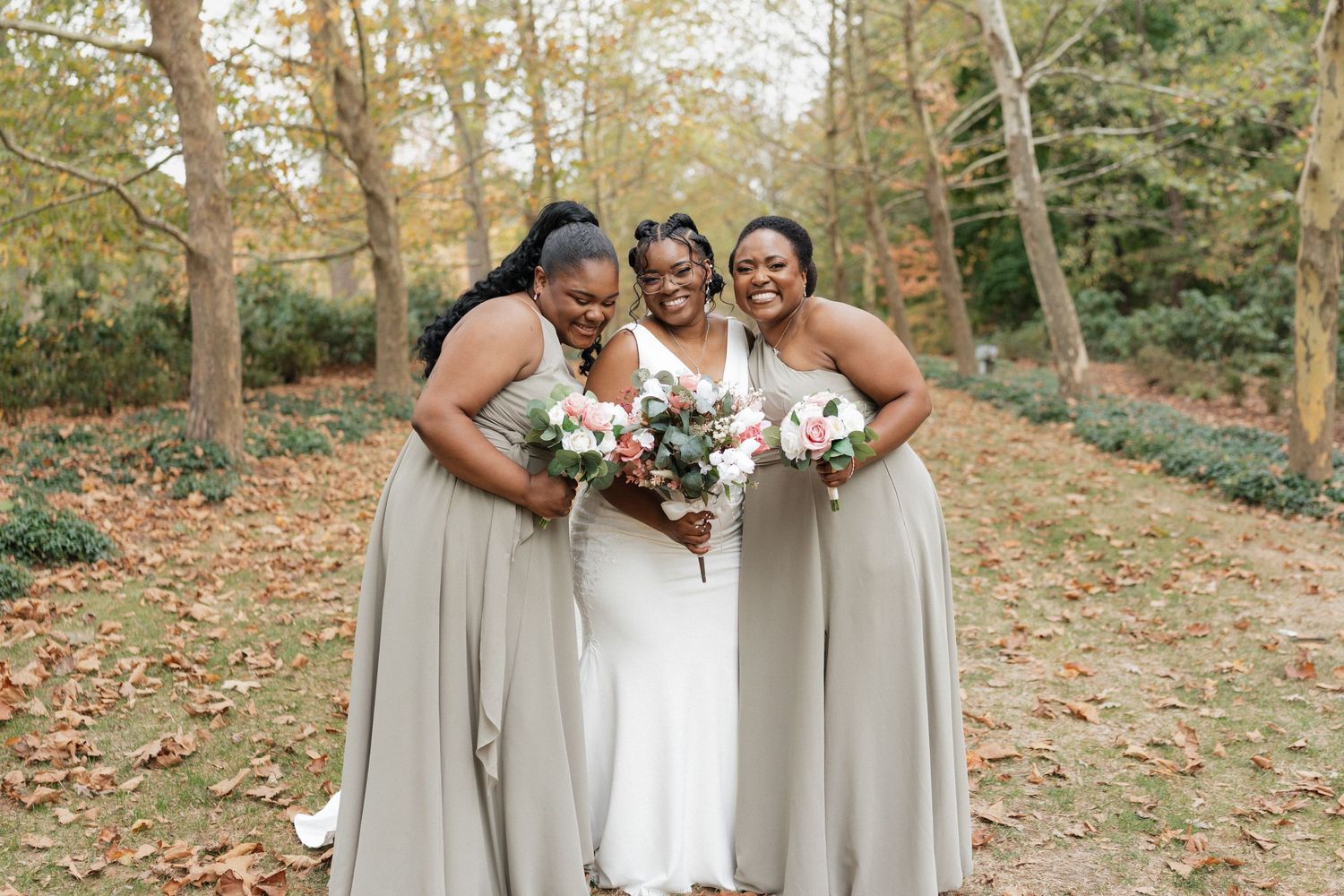 Bridesmaids in neutral dresses pose with floral bouquets against an autumn landscape.