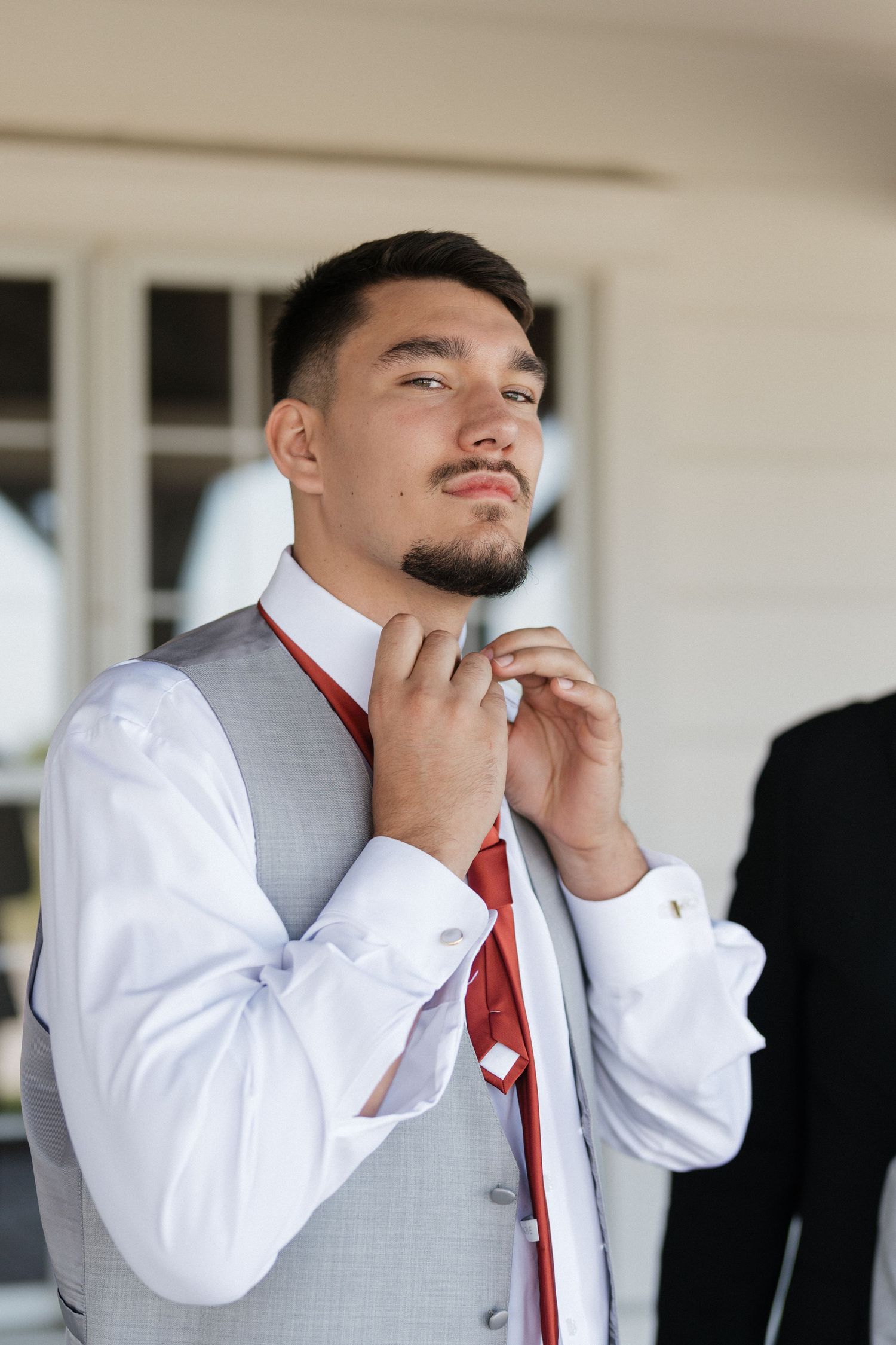 Groom in white shirt and gray vest adjusts his tie while getting ready for the wedding ceremony.