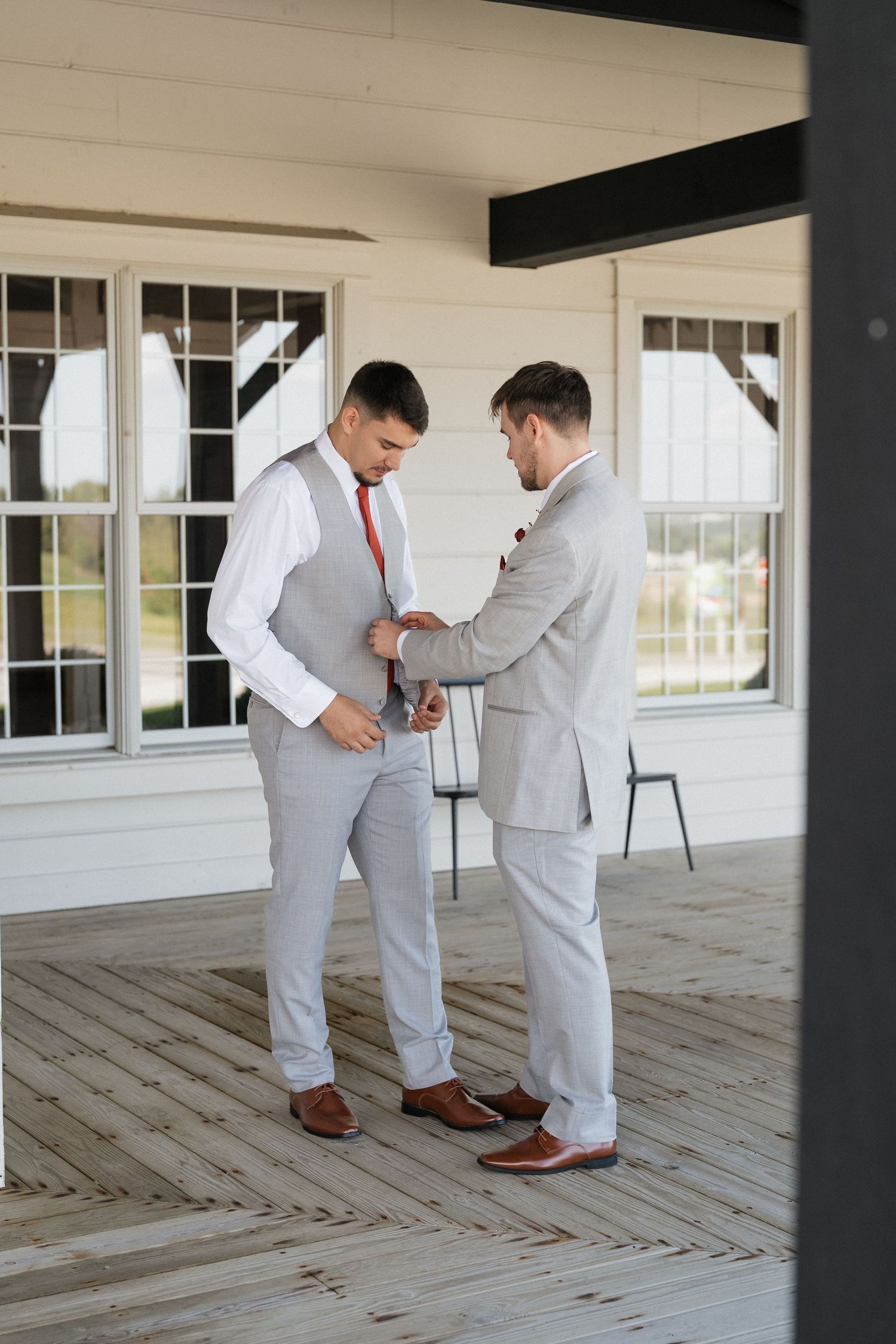 Two people in light gray suits helping each other get ready on a white porch.