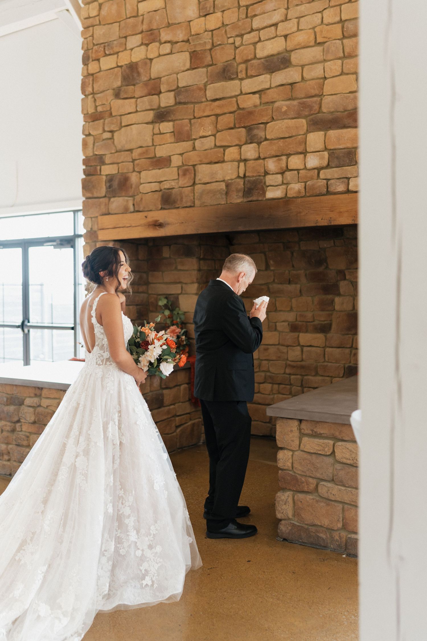 A bride in a lace gown stands near a stone fireplace during a first look moment.