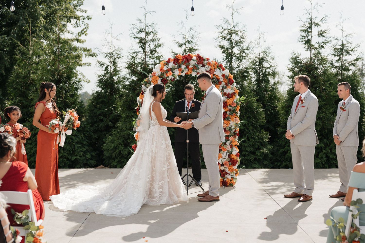 Wedding ceremony setup outdoors with floral arch and guests seated on white chairs under trees.