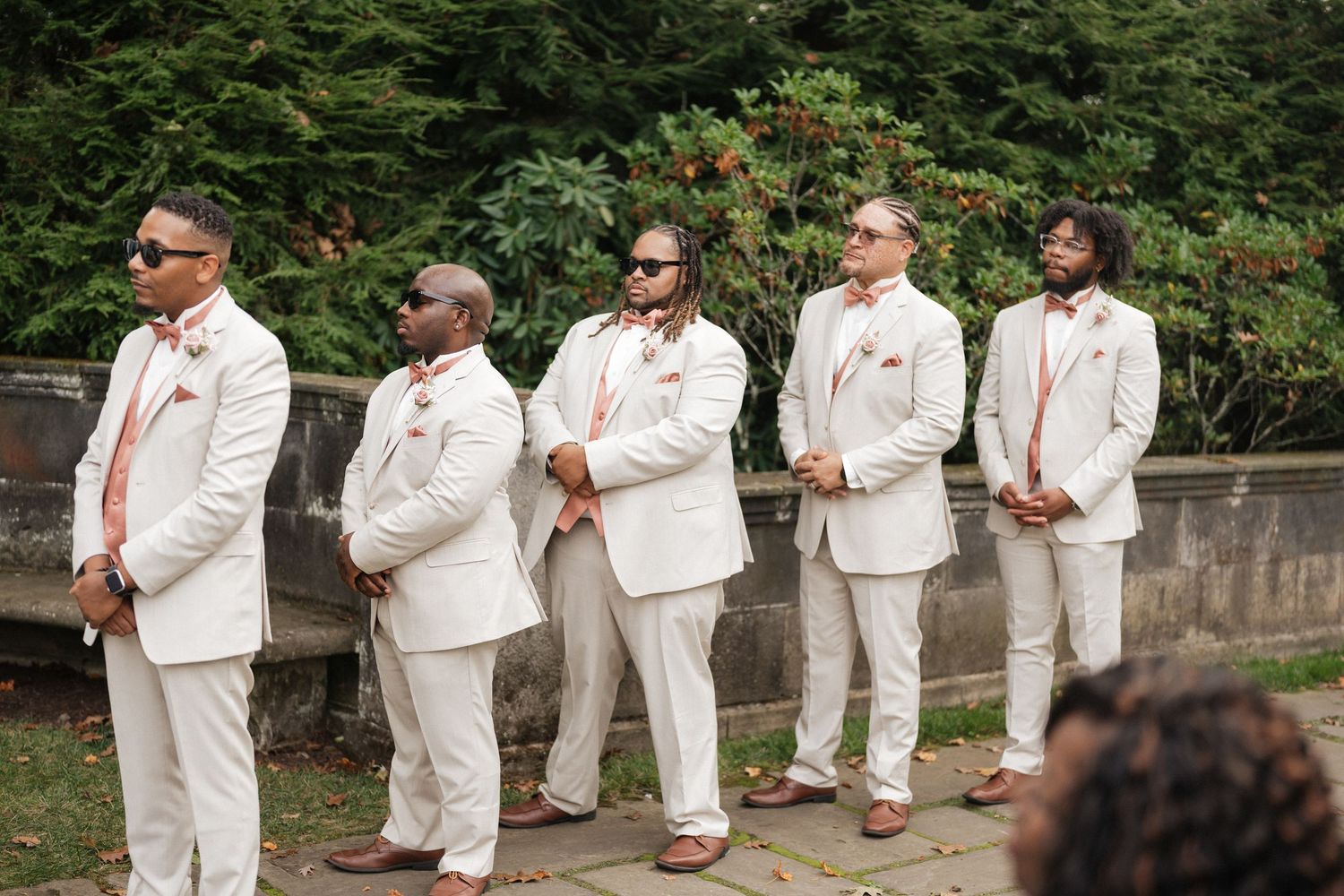 Groomsmen in white suits stand in line against garden backdrop.