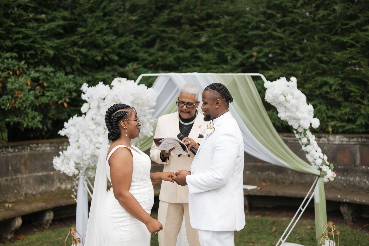 A wedding ceremony takes place outdoors under a white floral arch with lush green trees in the background.