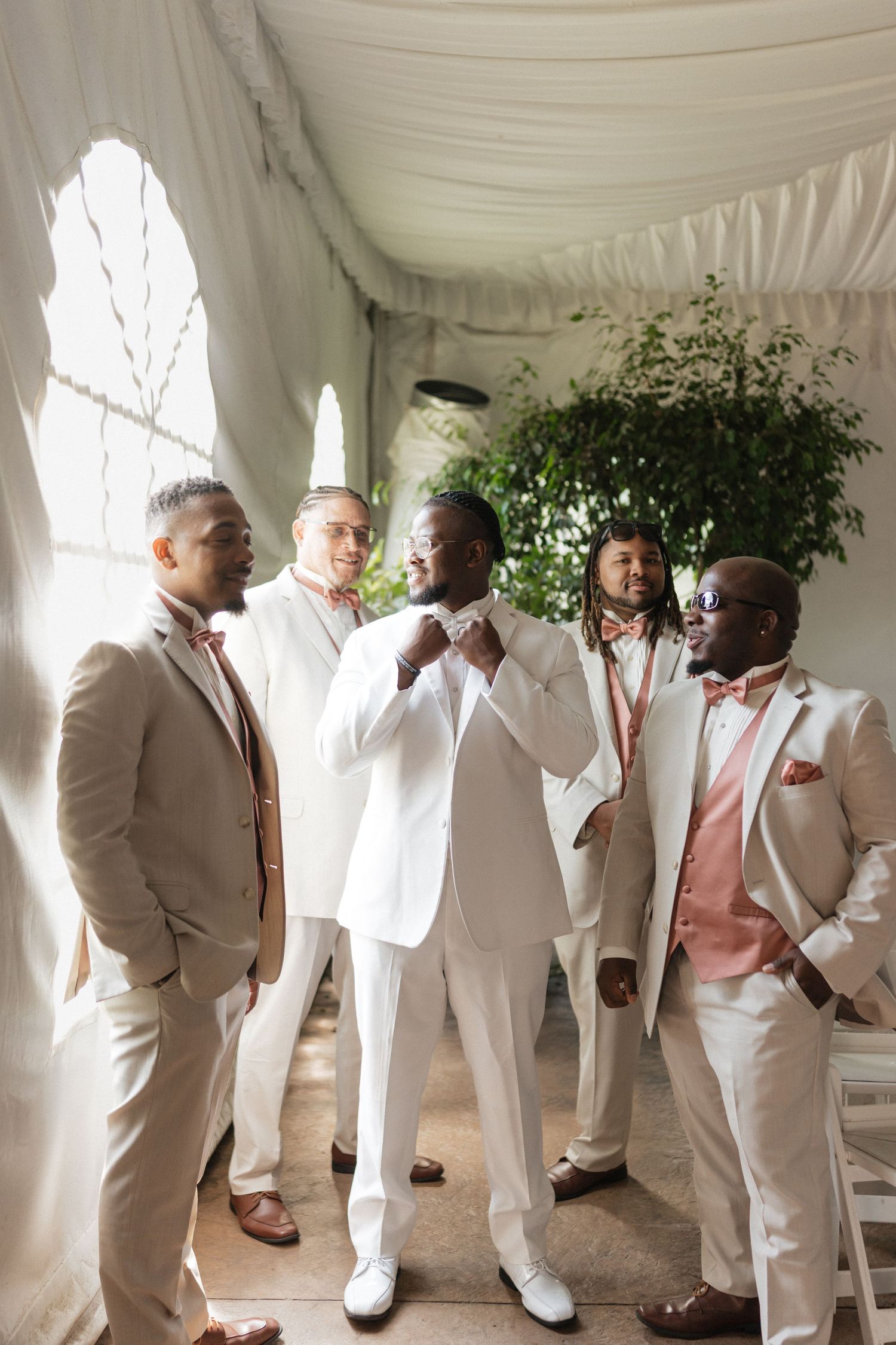 Groomsmen in crisp white and beige suits pose together under an elegant white tent.