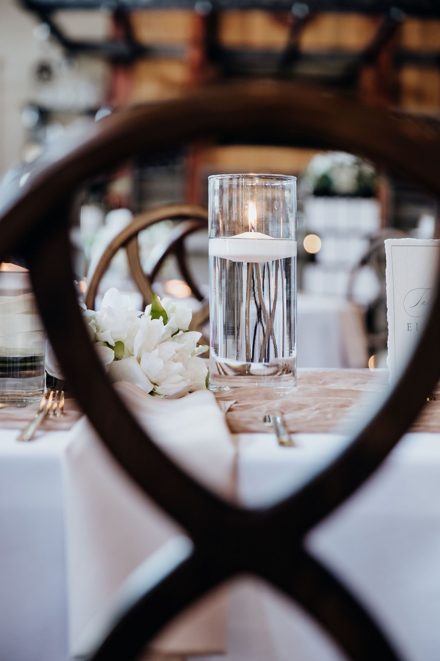 Wedding table settings with white floral centerpieces and glass votives captured through chair detail.