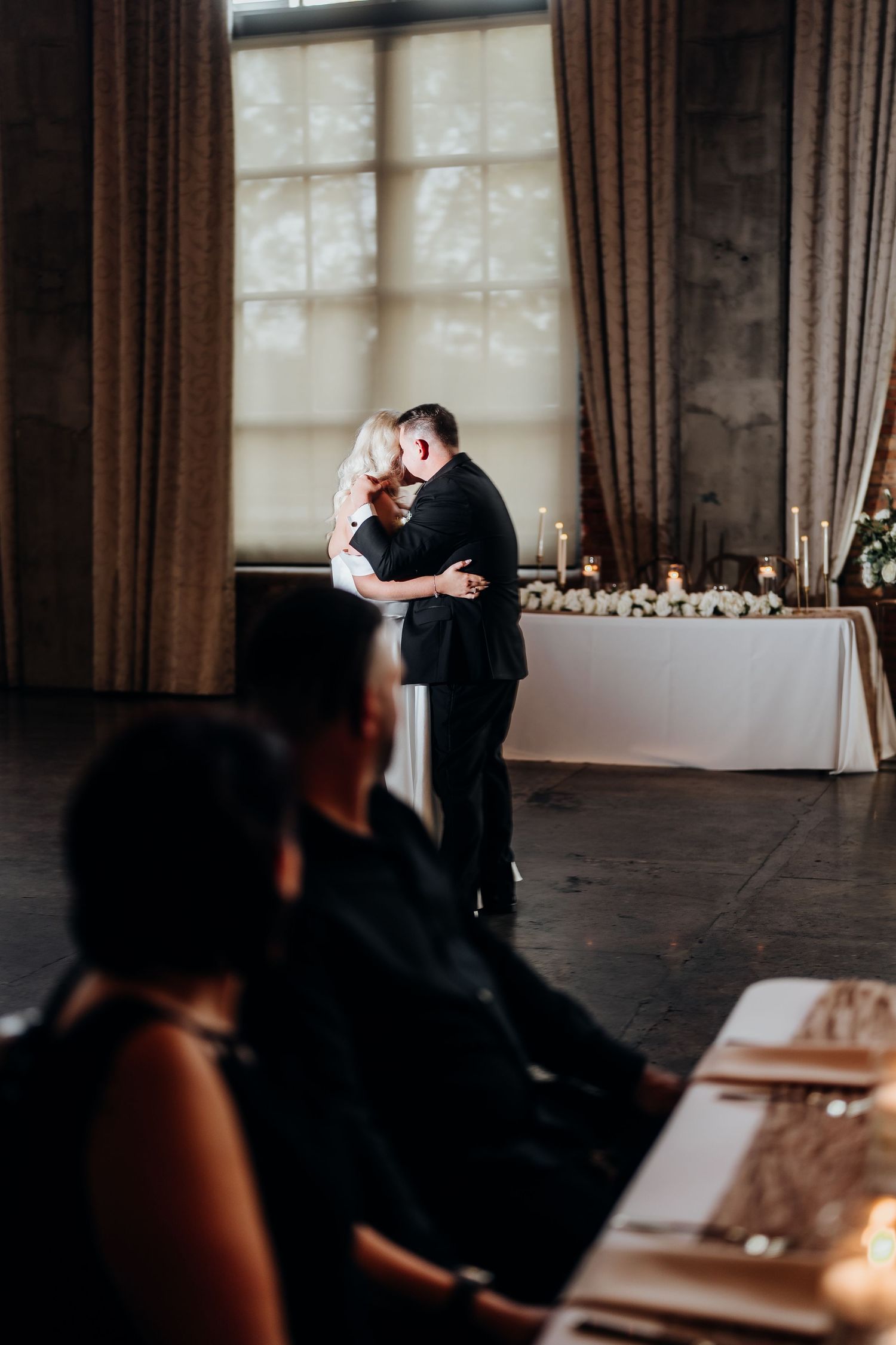 Couple sharing an intimate moment in a dimly lit reception venue with elegant window lighting.