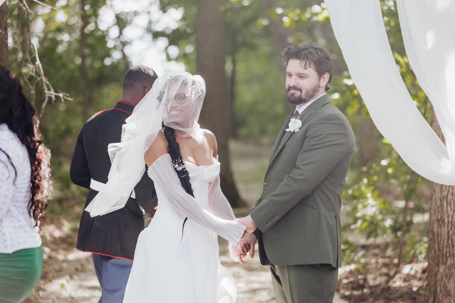 Wedding ceremony guests gathered outdoors among trees with white chairs.