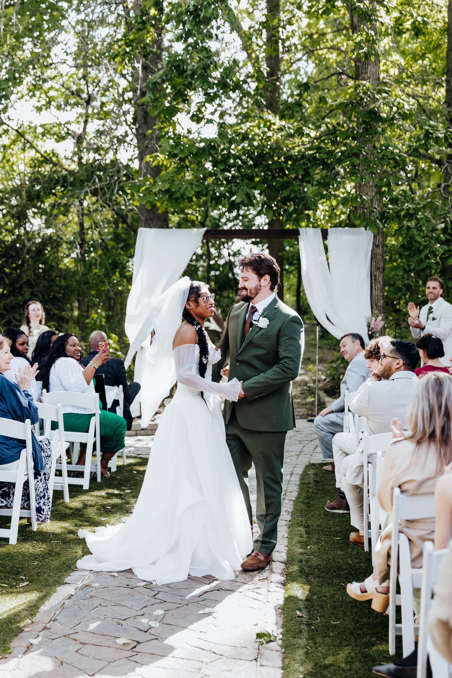 Outdoor wedding ceremony with white chairs and fabric drapes between trees as guests gather on a stone path.