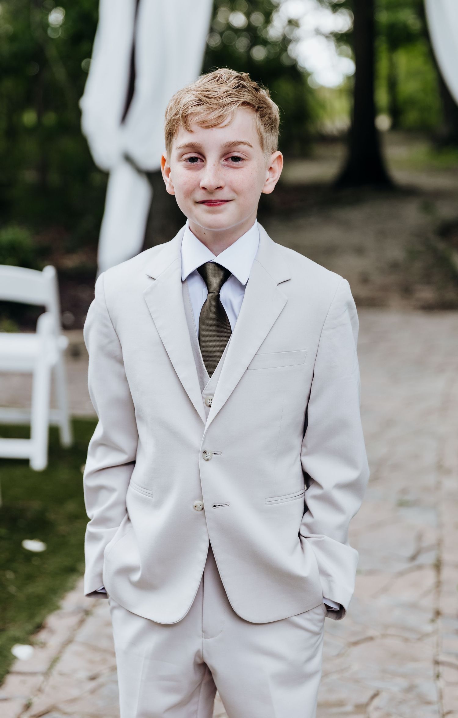 Young attendant in a crisp white suit poses with hands in pockets during wedding ceremony.