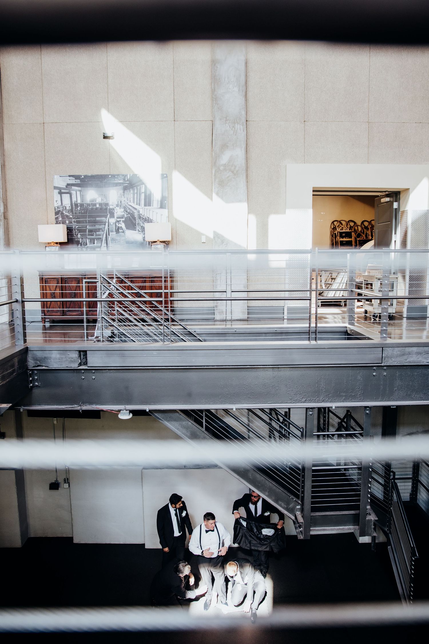 Groomsmen in black suits gather in an industrial space with white railings and concrete walls.