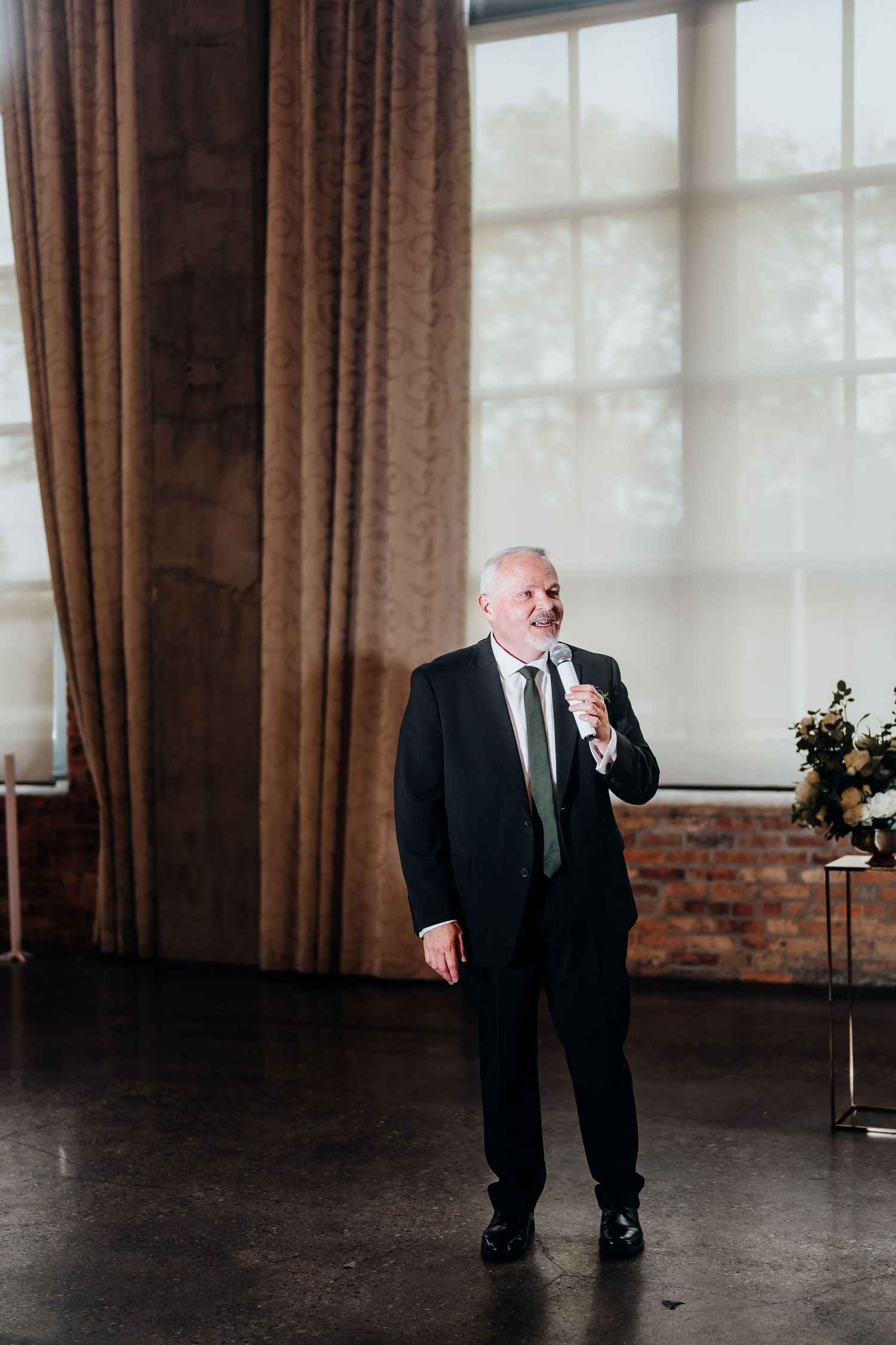 Man in black suit gives speech at elegant wedding venue with tall windows and curtains.