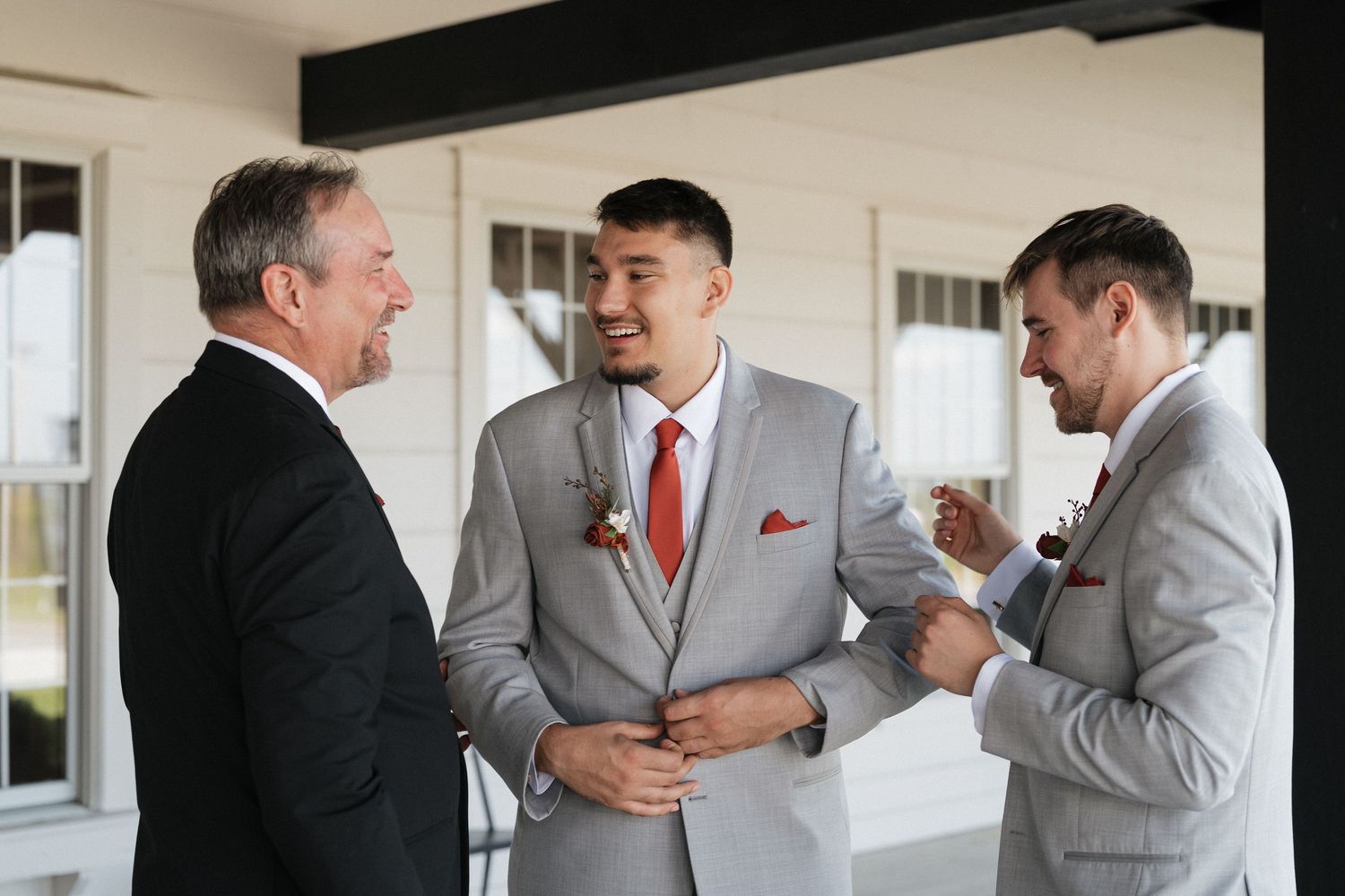 Wedding party in coordinated gray suits and red ties laughing together on a porch.