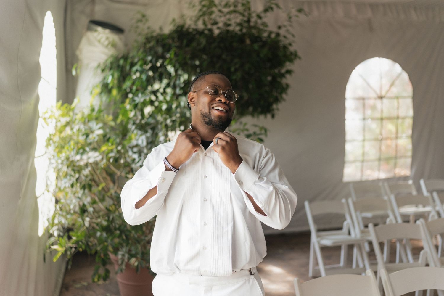 Someone adjusts a white bowtie while getting ready in an elegant white venue with arched windows and folding chairs.