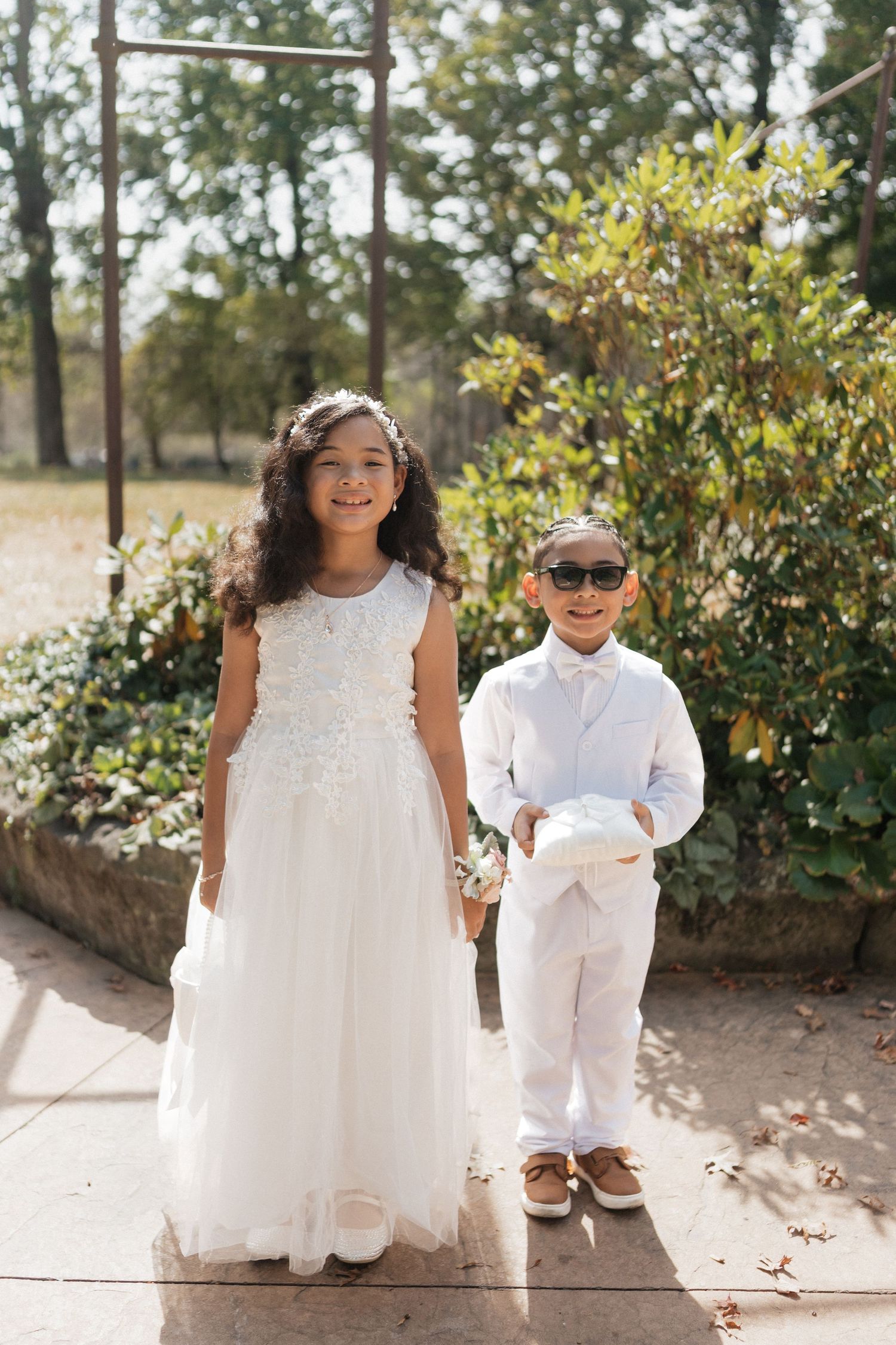 Children in white formal attire pose together outdoors against a sunny garden backdrop.