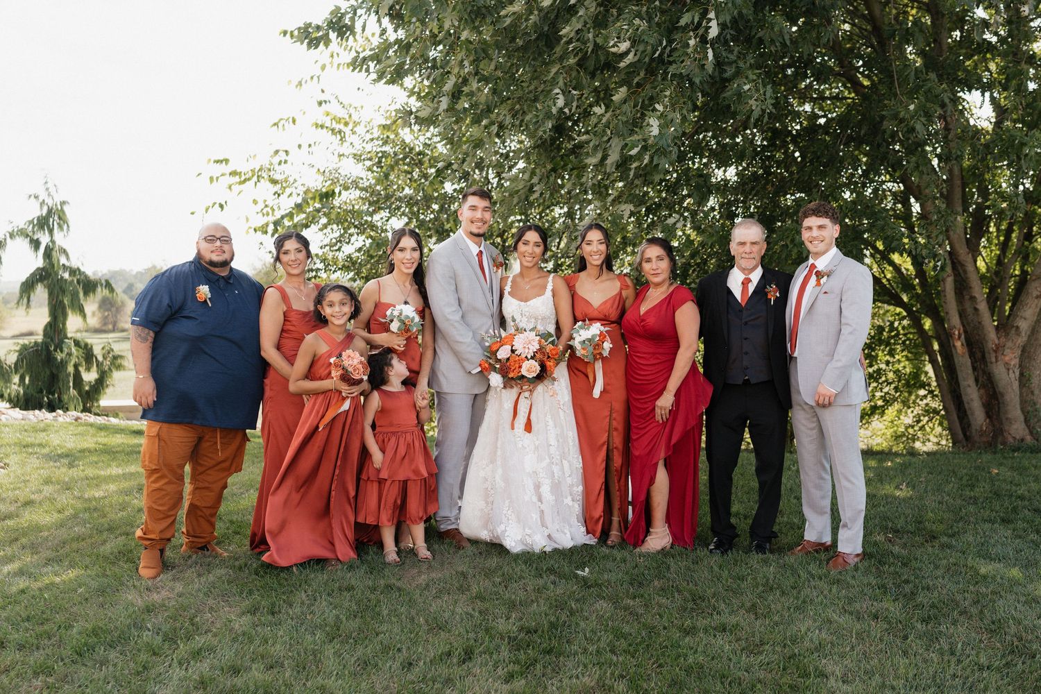 Wedding party in red and gray attire poses together on grassy lawn.
