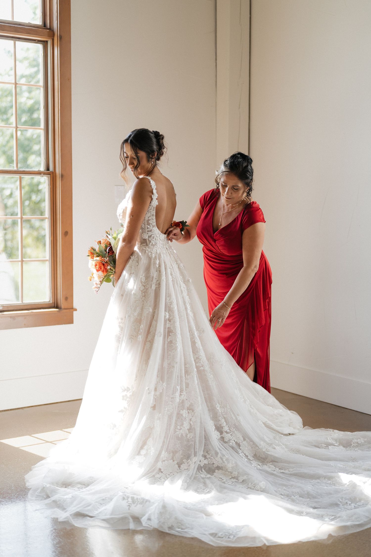 A bride in a lace wedding gown with long train gets help adjusting her dress near a sunlit window.
