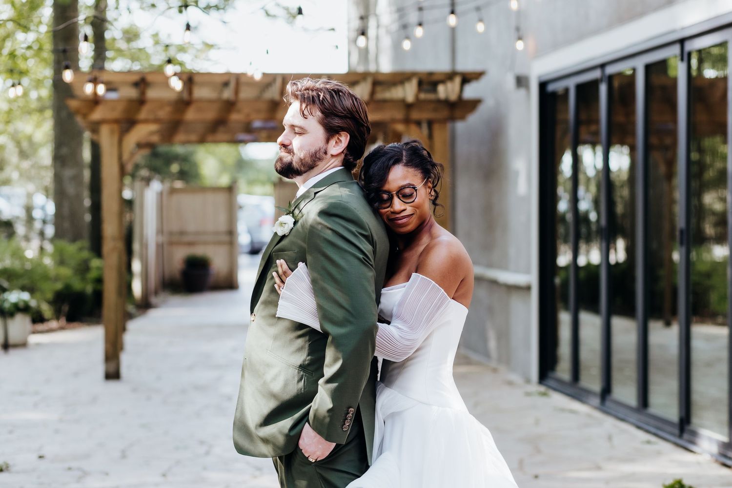 Couple poses back-to-back in green suit and white wedding gown outside modern venue.