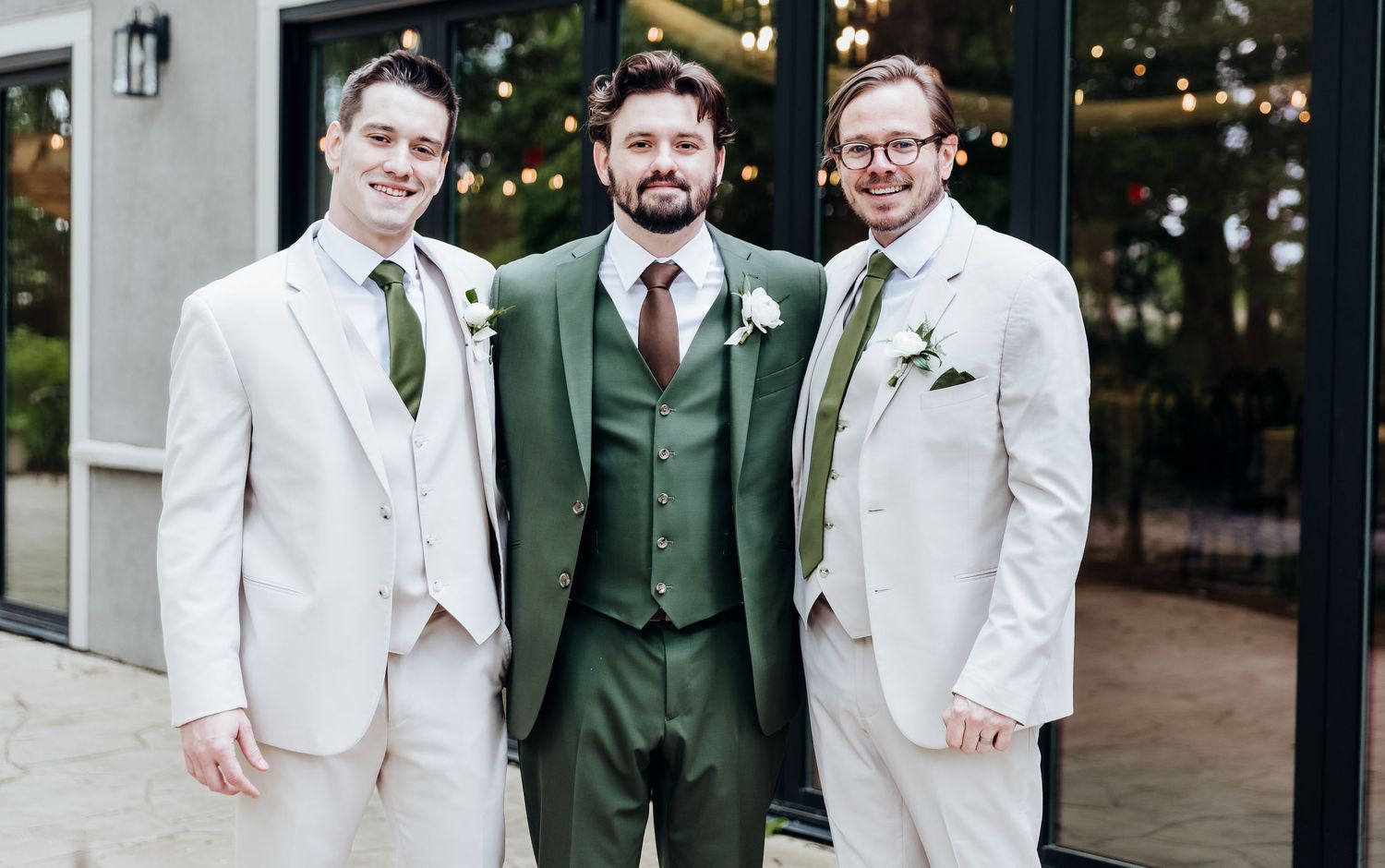 Three groomsmen pose together in contrasting white and dark green suits at an outdoor wedding venue.